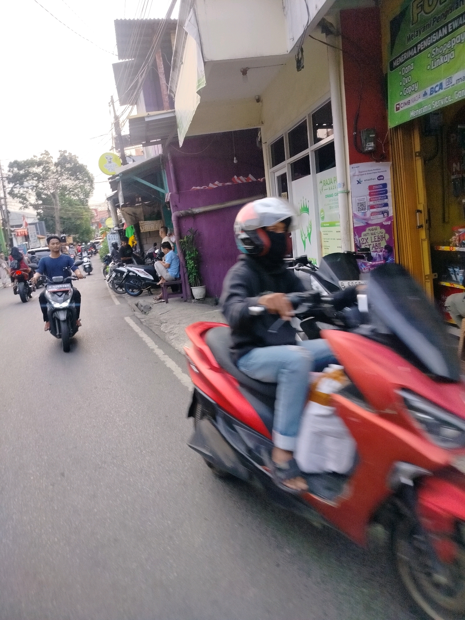 The image depicts a street scene in East Jakarta, Indonesia, possibly during the late afternoon or early evening due to the ambient light. In the foreground, a person wearing a helmet and a black mask is riding a red scooter, moving from left to right across the frame. This scooter is in motion, indicated by motion blur. The rider is wearing a black jacket and light blue jeans.
Further down the street, another person, a young man wearing a blue long-sleeved shirt and dark pants, is riding a black scooter. He is looking forward and appears to be traveling in the same direction as the foreground scooter. Behind him, several other motorcycles are parked or in motion.
The street is lined with buildings. On the right side of the image, a shop with yellow and red signage is visible, displaying text and advertisements. One sign on the right appears to list payment services. To the left of the shop, there is a building with a purple facade. Further down the street on the left, more buildings and trees are present. Power lines are strung across the scene overhead. The road surface is asphalt, with white lane markings visible. The overall atmosphere suggests a typical urban environment with active street traffic.