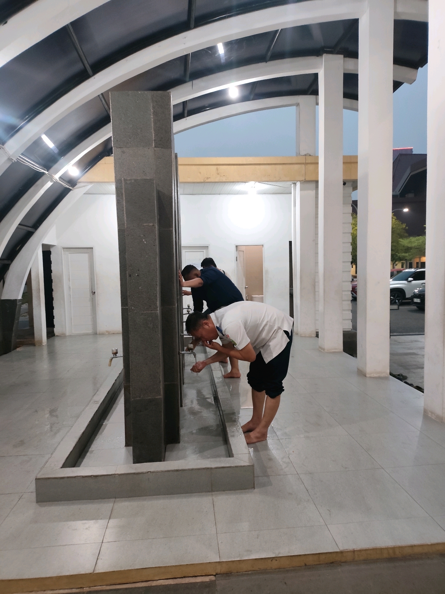 The image depicts an outdoor ablution area, likely at a mosque or religious facility, in Palembang, Indonesia. Three individuals are visible performing ablution, a ritual washing before prayer. In the foreground, a person wearing a white t-shirt and dark pants bends over a tap, cupping water to their face. Behind them, two other individuals, one in a dark blue shirt and the other in a dark t-shirt, are also engaged in the same activity. The ablution area features a row of dark stone pillars with water taps and a tiled floor that appears wet. The architecture includes a curved roof structure supported by white pillars, and in the background, white doors are visible on the left. The environment suggests it is either dusk or early morning, with outdoor lighting illuminating the scene. A street with vehicles, including a white SUV, is visible beyond the covered area.