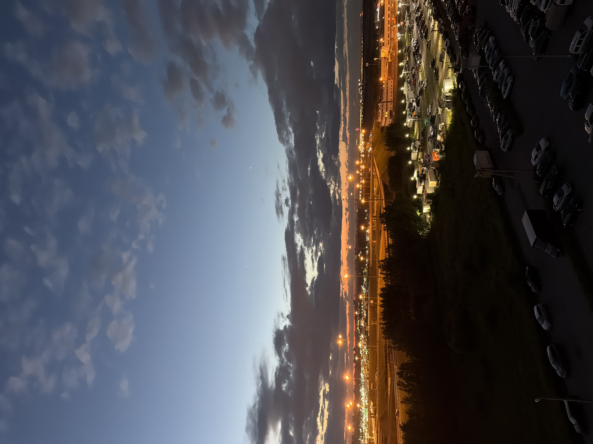 An aerial shot captures a dusk scene over a city, featuring a vast expanse of clouds against a gradient sky of deep blue to orange. In the mid-ground, a network of illuminated highways and roads winds through a landscape dotted with buildings and a large, brightly lit parking lot. Numerous cars fill the parking area, and streetlights cast a warm glow. The foreground is dominated by dark, silhouetted trees and a dimly lit area with several parked cars. The image appears to be taken from a high vantage point, offering a panoramic view of the urban environment as day transitions into night.