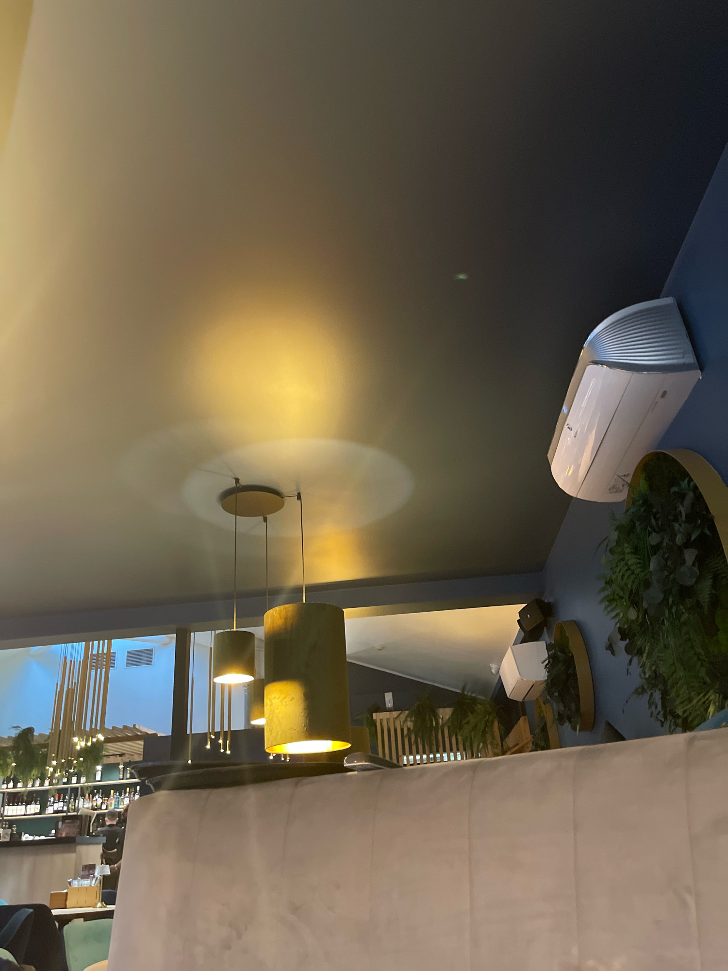 The image depicts the interior of a modern establishment, likely a restaurant or bar, with a focus on the ceiling and upper wall space. A dark grey ceiling dominates the upper portion of the frame, with a large, soft yellow light emanating from an unseen source, creating a diffuse glow and casting shadows. Two pendant lights, each with a cylindrical, olive-green lampshade, hang from the ceiling. An air conditioning unit, white with silver accents, is mounted on a deep blue wall in the upper right corner. To the left of the air conditioning unit, on the dark grey ceiling, a small green light is visible. The lower portion of the image reveals a plush, light-colored upholstered seating area, likely a sofa or booth, suggesting a comfortable seating arrangement for patrons. In the background, through a large window or open space, a bar area is visible with shelves stocked with bottles and a few vertical wooden decorative elements. The overall ambiance appears to be dimly lit and sophisticated. Based on the visual cues, such as the modern decor and lighting, this establishment is likely situated in a contemporary urban setting. Without further information or context, it is not possible to definitively pinpoint the location within Tallinn, Estonia.