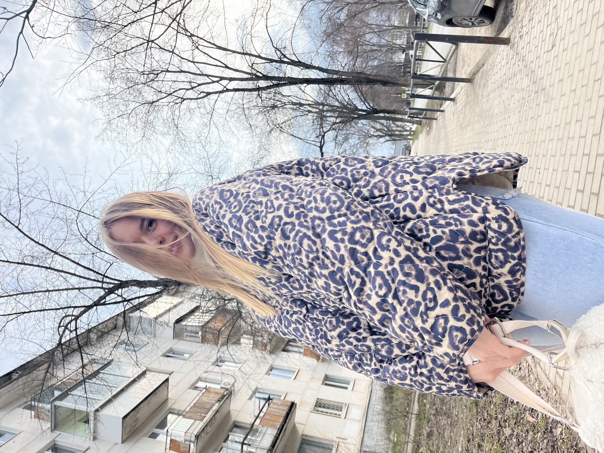 The image shows a woman standing on a brick sidewalk, likely in an urban area. She is wearing a leopard-print puffer jacket and light blue jeans, and she is holding a white tote bag with a light-colored strap. Behind her is a residential building with visible balconies and windows. Above the building are bare tree branches against a cloudy sky. In the distance, a street with a black fence, a sidewalk and a car can be seen. Based on location data, the setting is within the city of Chaikovsky, Russia.