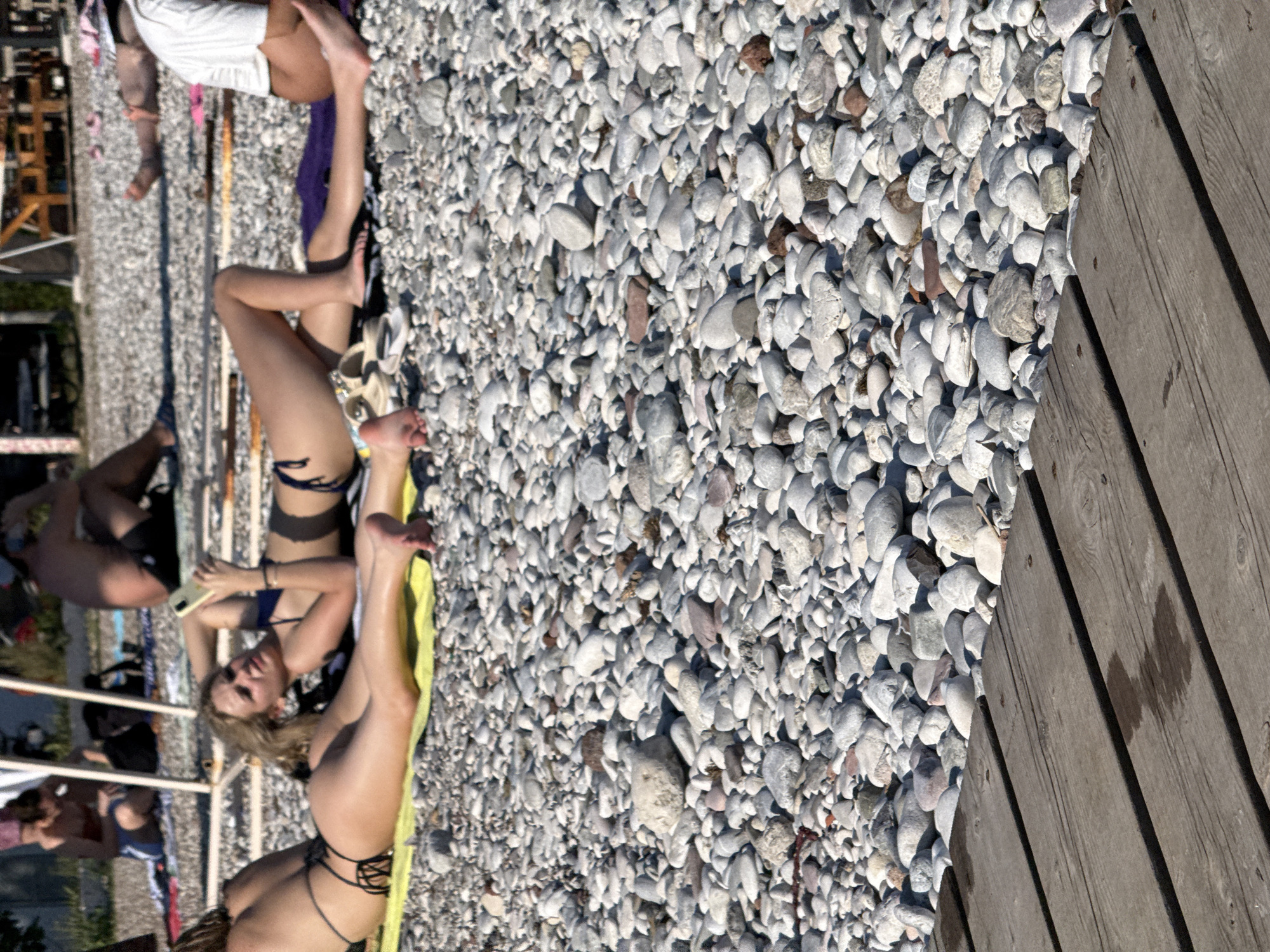 The image shows a group of people on a pebble beach in Budva, Montenegro. The sun is shining brightly, casting shadows on the beach, indicating it is daytime. The people are dressed in swimwear, suggesting they are on vacation or enjoying a day at the beach. One woman is lying on a yellow towel, looking at the camera and holding a phone. Another woman is lying on her side, with her legs extended. Other people are visible in the background, some sitting and others lying down. The beach is covered in small, smooth pebbles, and a wooden boardwalk is visible on the right side of the image. The overall atmosphere appears relaxed and leisurely, consistent with a beach setting. No text is visible in the image.