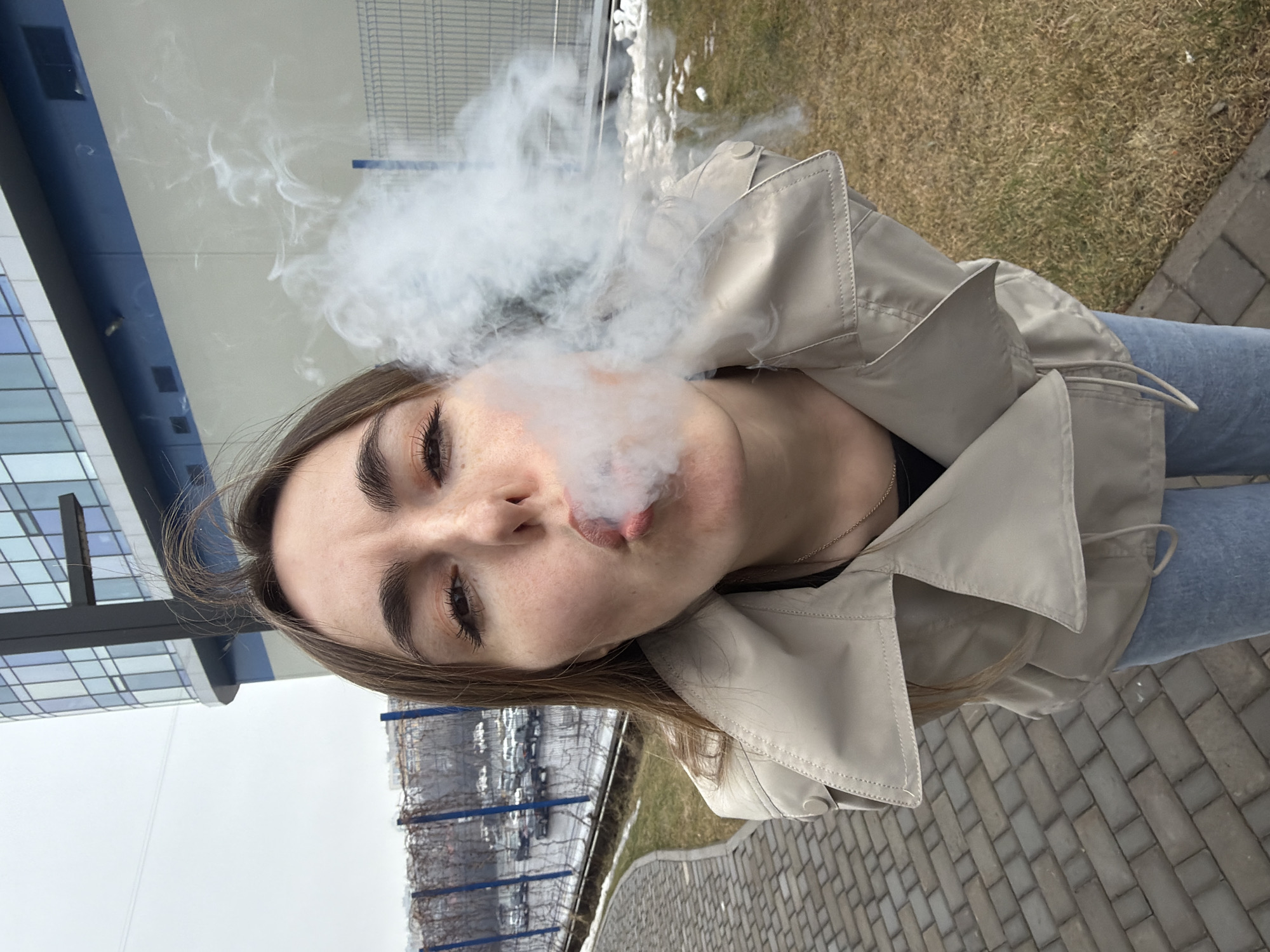 A woman is pictured from a low-angle perspective, facing the camera while exhaling a large cloud of white vapor or smoke from her mouth. The background consists of urban elements: a building with a blue and white façade, a fenced area with bare trees, a patch of dry grass, and a paved surface. The location is Izhevsk, Russia. The woman is wearing a tan trench coat and jeans. She has light brown hair and her face is turned slightly to the side.