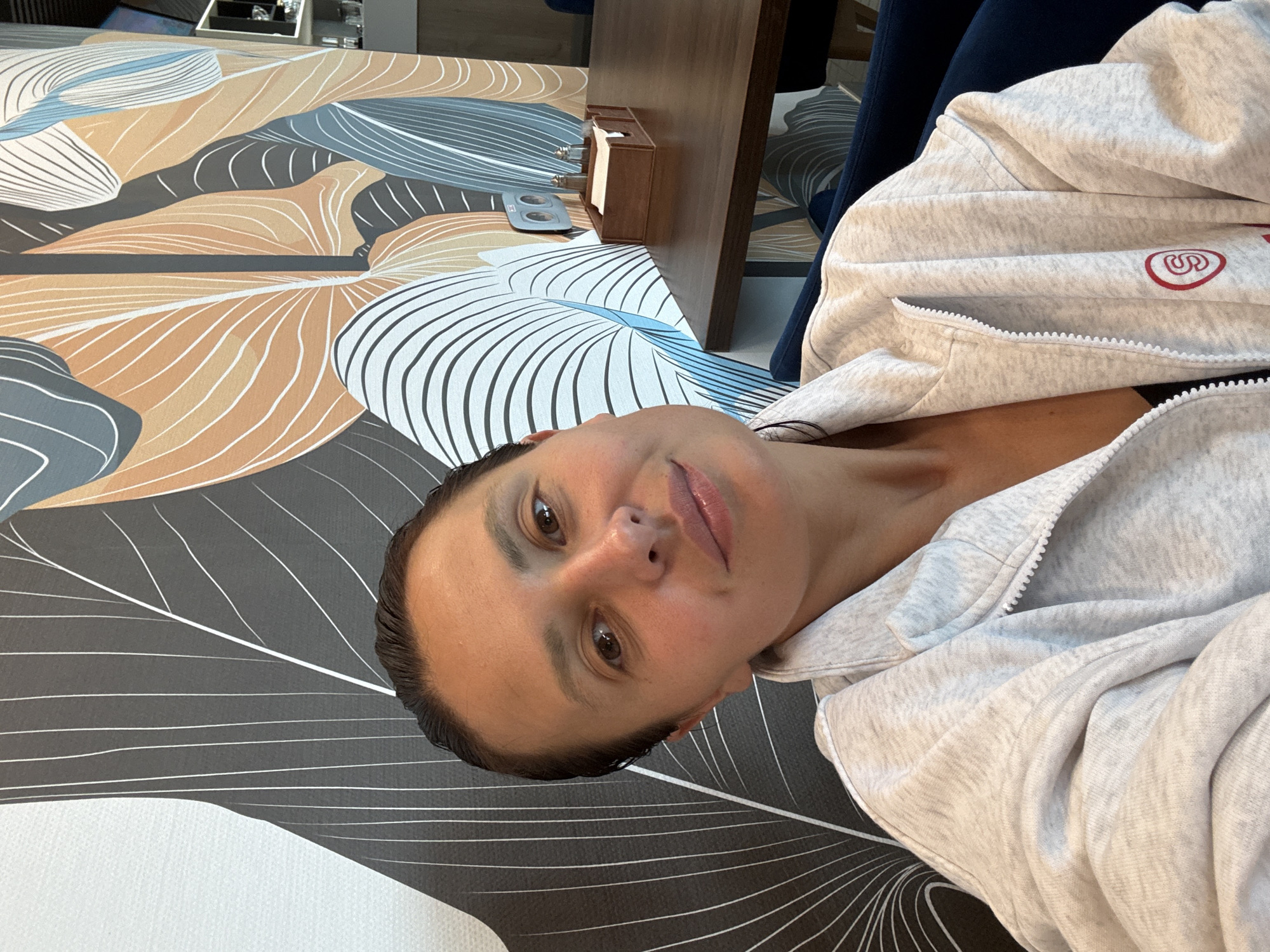 The image captures an adult female taking a self-portrait within an indoor commercial establishment located in Chaikovsky, Russia. The woman, positioned centrally, looks directly into the camera with a subtle smile and a slight tilt of her head to the viewer's left. Her dark hair is pulled back and appears sleek. She is wearing a light grey heathered zip-up jacket with a white zipper, under which a dark garment is visible. On the wearer's left chest, there is a circular red embroidered logo containing a stylized red 'S'.
The background consists of a wall covered in a distinct large-scale botanical patterned wallpaper featuring stylized leaves and veins in shades of dark grey, white, light orange, and light blue. To the woman's right, a dark brown wooden table is visible, upon which sits a wooden caddy containing white paper napkins and two condiment shakers with silver tops. A section of a dark blue upholstered chair or booth is visible beneath the woman and on the far side of the table. A white double electrical socket is mounted on the patterned wall above the table. Further in the background, dimly lit shelves and a dark screen are partially visible.