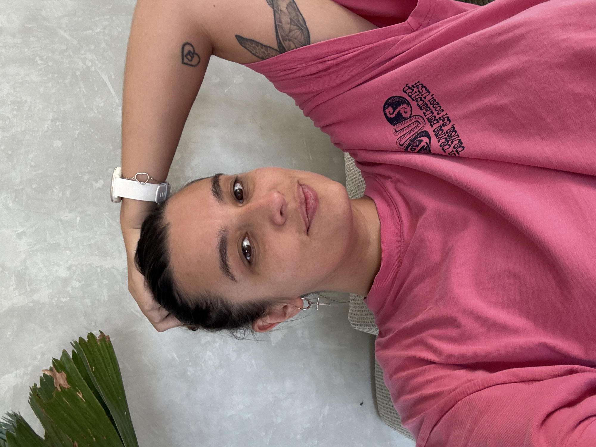 A woman with dark hair pulled back is wearing a pink t-shirt with a snake logo and text on the sleeve, a white watch with a heart-shaped accent on the band, and small silver cross earrings. She has tattoos on her left arm; a small heart-shaped outline on her forearm and a larger tattoo depicting a bird on her upper arm. The background consists of a textured grey wall with a green plant visible on the left side of the frame. Based on the text on the shirt, the location of the image is in the vicinity of or associated with Ubud District, Indonesia. The woman is in a seated position with her arm bent upward.