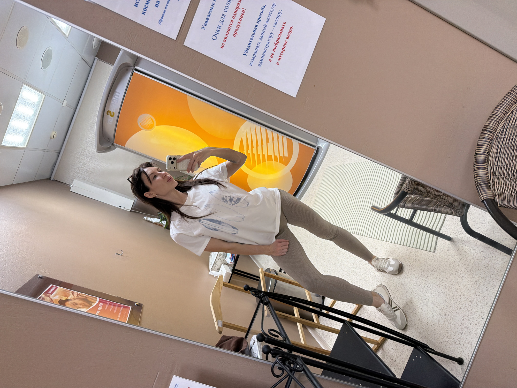 The image shows a woman taking a mirror selfie in what appears to be a solarium or tanning studio. The woman is wearing a white t-shirt, leggings, and sneakers. She is holding her phone up to take the photo. The background includes tanning equipment, information signs in Russian, and furniture. Based on the Russian signage, it could be located in Perm, Russia.