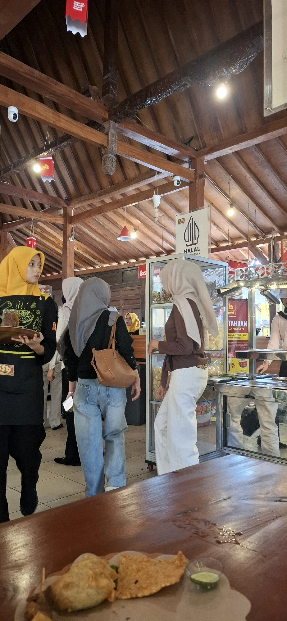 A vertical, medium shot captures the interior of a restaurant in Colomadu, Indonesia, characterized by its wooden architecture and a bustling atmosphere. The scene is populated by several individuals, most wearing hijabs. In the foreground, a wooden table bears a plate of food, including what appears to be fried snacks and a slice of lime.
To the left, a person wearing a yellow hijab and a black apron with designs stands, holding a tray with a drink. Behind them, another person in a grey hijab is facing away, holding a brown purse. Further back, a person in a light brown hijab is examining items in a display case.
The restaurant's ceiling features exposed wooden beams and traditional carvings. Several red and white banners with Indonesian text hang from the ceiling, suggesting a festive or nationalistic theme. Two security cameras are visible mounted on the wooden beams.
The display case, partially visible, contains various food items and promotional materials. A sign with the word "HALAL" in Arabic script and English is prominently displayed above the case. The flooring is tiled. The overall impression is of a lively dining establishment with a cultural ambiance.