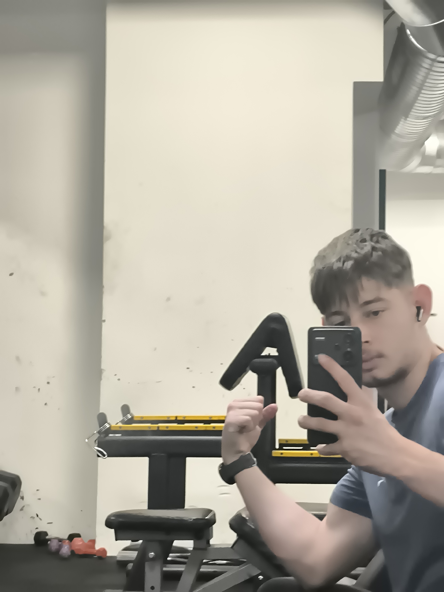 A person is taking a selfie in a gym. The gym appears to be in an interior space with light-colored walls and a visible ventilation system in the upper right corner. In the foreground, there is gym equipment including an arm curl machine. On the floor, there are some small weights. The person in the image is holding a smartphone in their right hand to take the selfie, showing their arm flexing. They are wearing a smartwatch on their left wrist and an earbud. The location cannot be determined.
