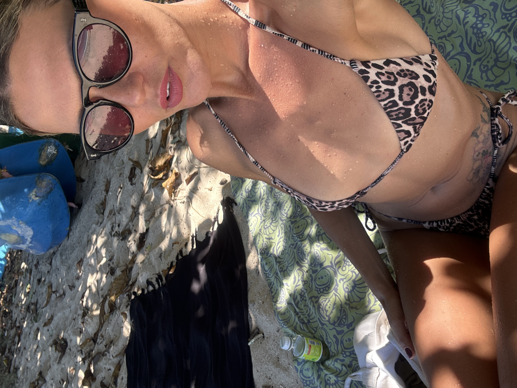 A person is seen lying outdoors on a sunny day, likely at a beach or sandy area in Kuta Selatan, Indonesia. The main subject, a person with wet, glistening skin, is wearing a leopard-print bikini and dark, round sunglasses. Their lips are slightly parted, revealing a hint of teeth and appear to have pink lipstick. A tattoo is partially visible on their left side, near the hip.
The setting appears to be a sun-drenched environment, with strong sunlight casting distinct shadows across the scene, suggesting midday or early afternoon. The person is partially resting on a light green towel adorned with a swirling, organic pattern. Around them, the ground consists of sand scattered with dry leaves.
Various objects are present in the scene: a dark-colored fabric item, possibly a towel or clothing, lies on the sand to the left of the person. A blue object, possibly a buoy or part of a small boat, is partially visible in the upper left corner of the image. Near the person's legs, on the green towel, there is a small, clear plastic bottle with a white cap and a yellow label, which has unreadable text. A crumpled white fabric item is also seen near their right leg. The overall activity suggests relaxation or sunbathing, possibly after a swim.