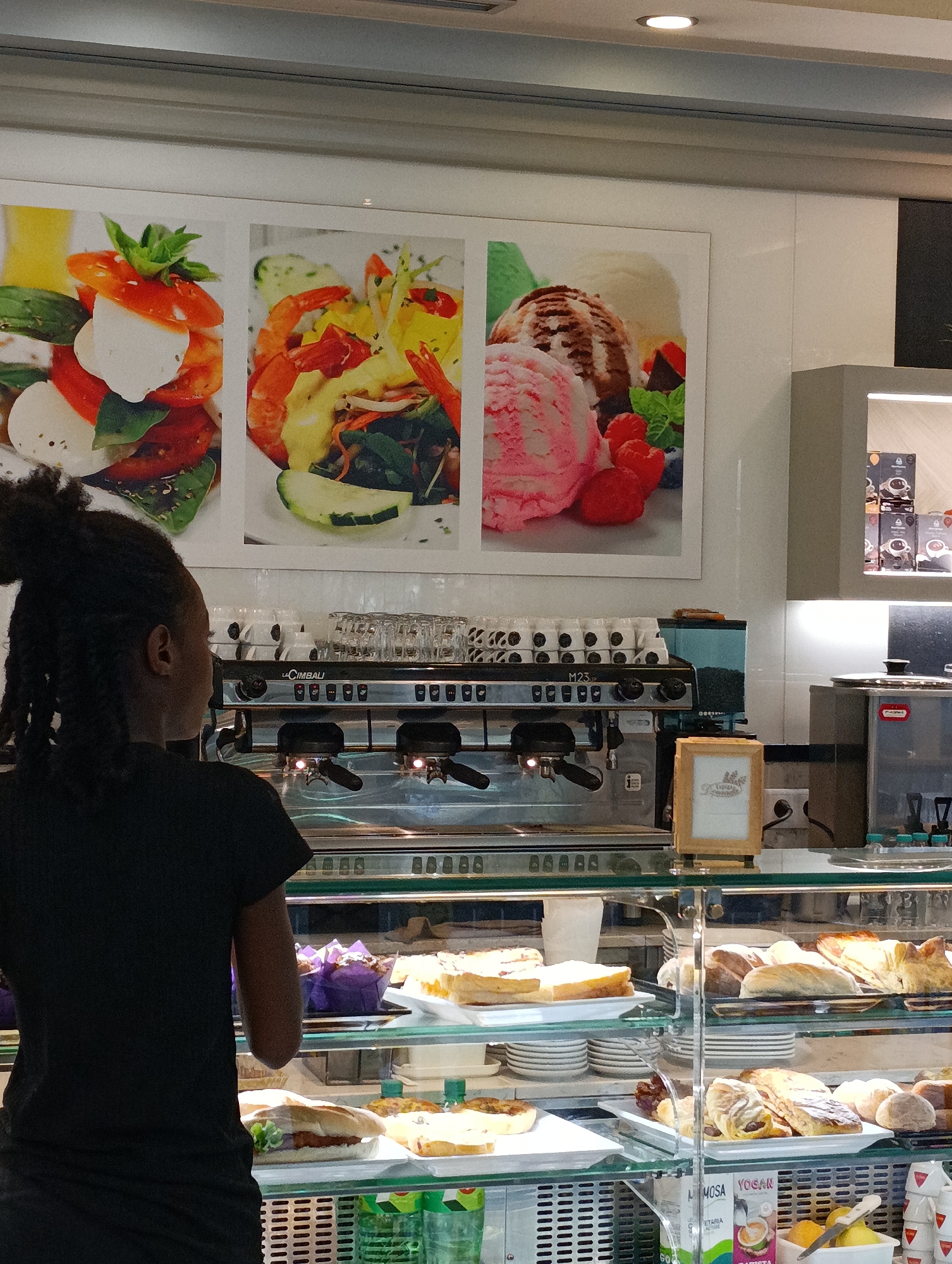 The image shows a young woman with dark hair, facing away from the camera, looking at a display of food items. She is wearing a black t-shirt. Behind her is a coffee machine with a display of cups stacked on top. Above the coffee machine are three framed pictures: one of a salad with tomatoes, mozzarella, and basil; another of a seafood salad with shrimp and vegetables; and a third of ice cream scoops with berries. In front of the woman is a glass display case filled with pastries, sandwiches, and drinks. The setting appears to be a cafe or bakery. There is a sign on a wooden stand in front of the display case. The lighting suggests it is daytime. Some visible text includes "La Cimbali" on the coffee machine and "YOGAN" and "MIMOSA" on a juice carton in the display case.