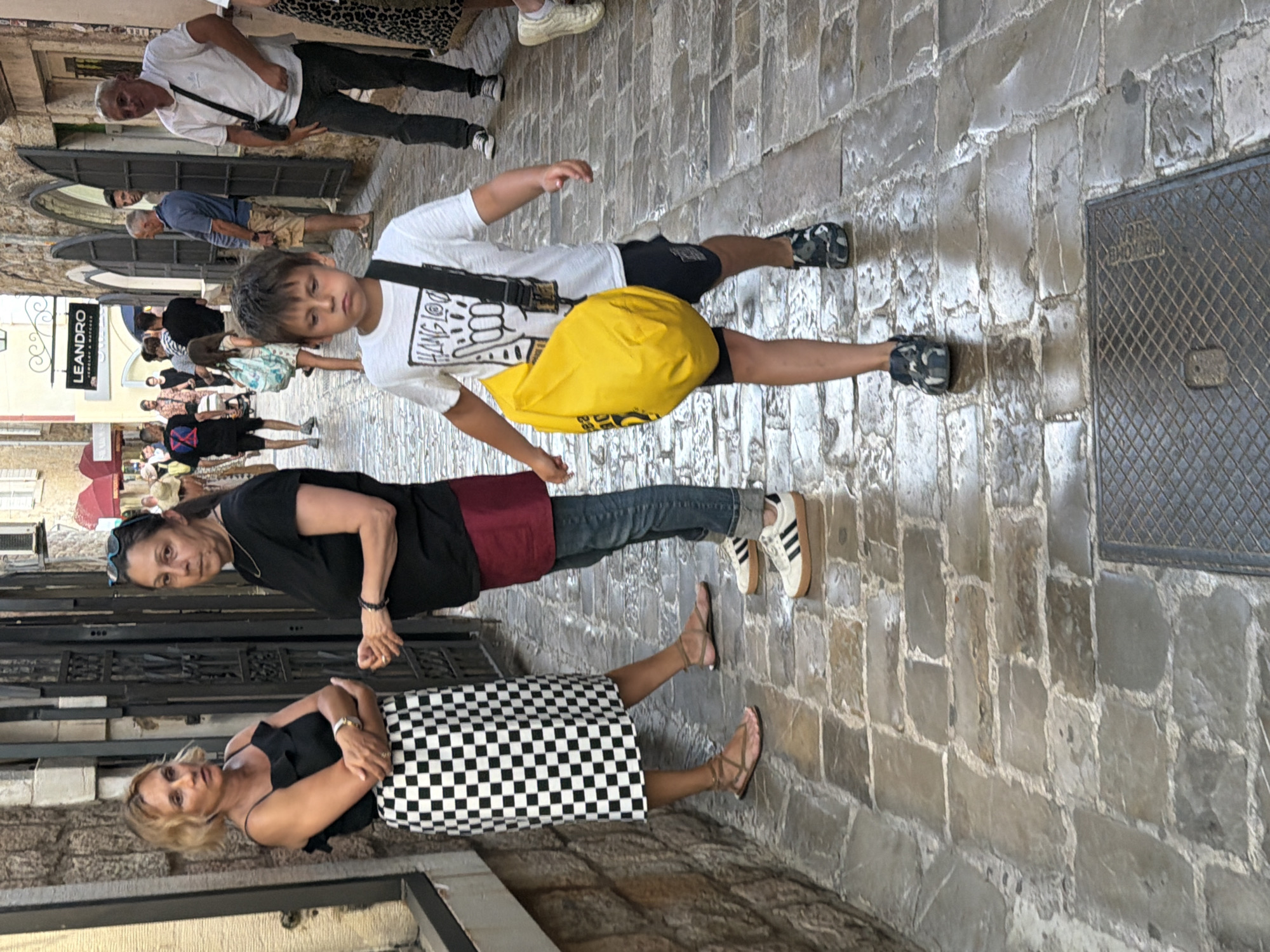 In the city of Budva, Montenegro, a cobblestone alleyway features a group of people. In the foreground, a young boy with dark hair walks towards the viewer, carrying a bright yellow bag slung across his body. He wears a white t-shirt and dark shorts with camouflage-patterned shoes. Behind him, two women stand talking. The woman on the left has blonde hair and wears a black top and a black and white checkered skirt with sandals. The woman on the right has dark hair and wears a black t-shirt and a burgundy skirt with jeans and white sneakers. In the background, several other individuals are visible walking through the alley, some standing and conversing. The alley is lined with stone buildings, one of which has a sign that reads "LEANDRO JEWELRY & WATCHES." The street is wet in patches, suggesting recent rain or cleaning.
