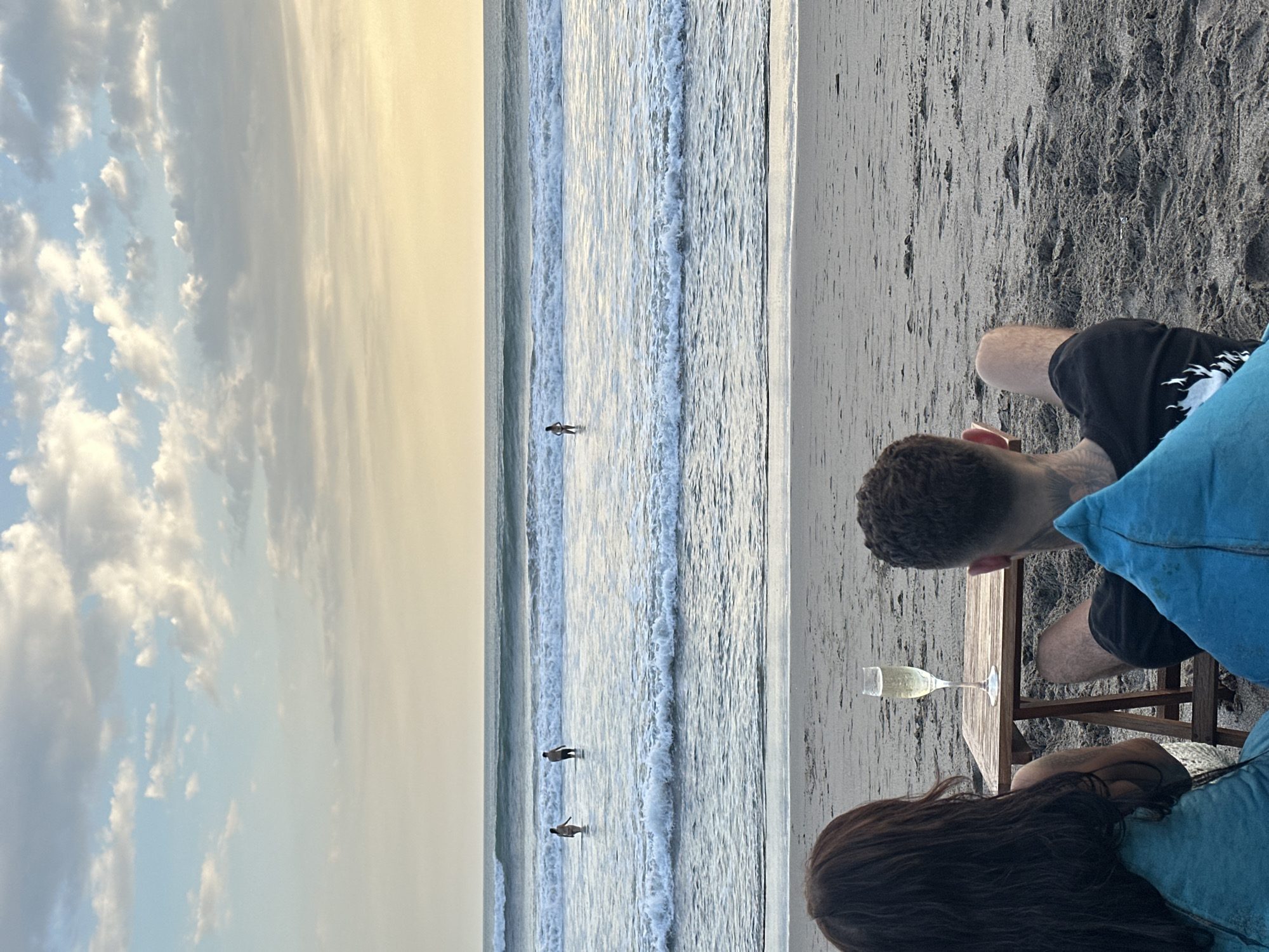A couple is seated at a low wooden table on a black sand beach, possibly in the North Kuta area of Indonesia, indicated by the beach type and ocean conditions. They appear to be enjoying a drink, as a glass of what seems to be white wine sits on the table. The ocean is calm with small waves, and several people are wading in the water. The sky is cloudy with a mix of gray and yellowish light, suggesting either sunset or sunrise.