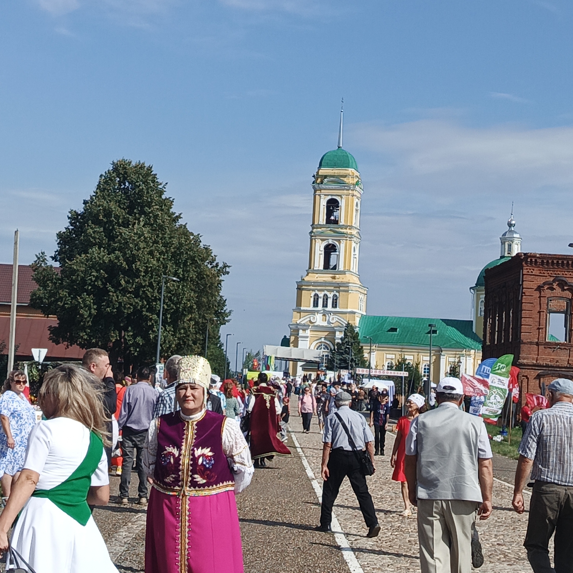 The image depicts a street scene in Nikolo-Beryozovka, Russia, with a clear blue sky overhead. A prominent yellow bell tower with a green dome and a slender spire rises in the background, attached to a church with a green roof. To the right of the bell tower, a brick building with arched windows is visible. In the foreground, a diverse crowd of people is walking on a cobblestone street. Several individuals are wearing traditional clothing, including a woman in the foreground on the left with a white and green dress and another woman on the right wearing a purple and gold ornate vest with a matching pink skirt and an elaborate headdress. Many people are dressed in casual attire. Flags, some with Cyrillic lettering, are displayed in the middle ground. Trees, including a large, leafy one on the left, and streetlights are also present in the environment. The scene suggests a public gathering or festival.
