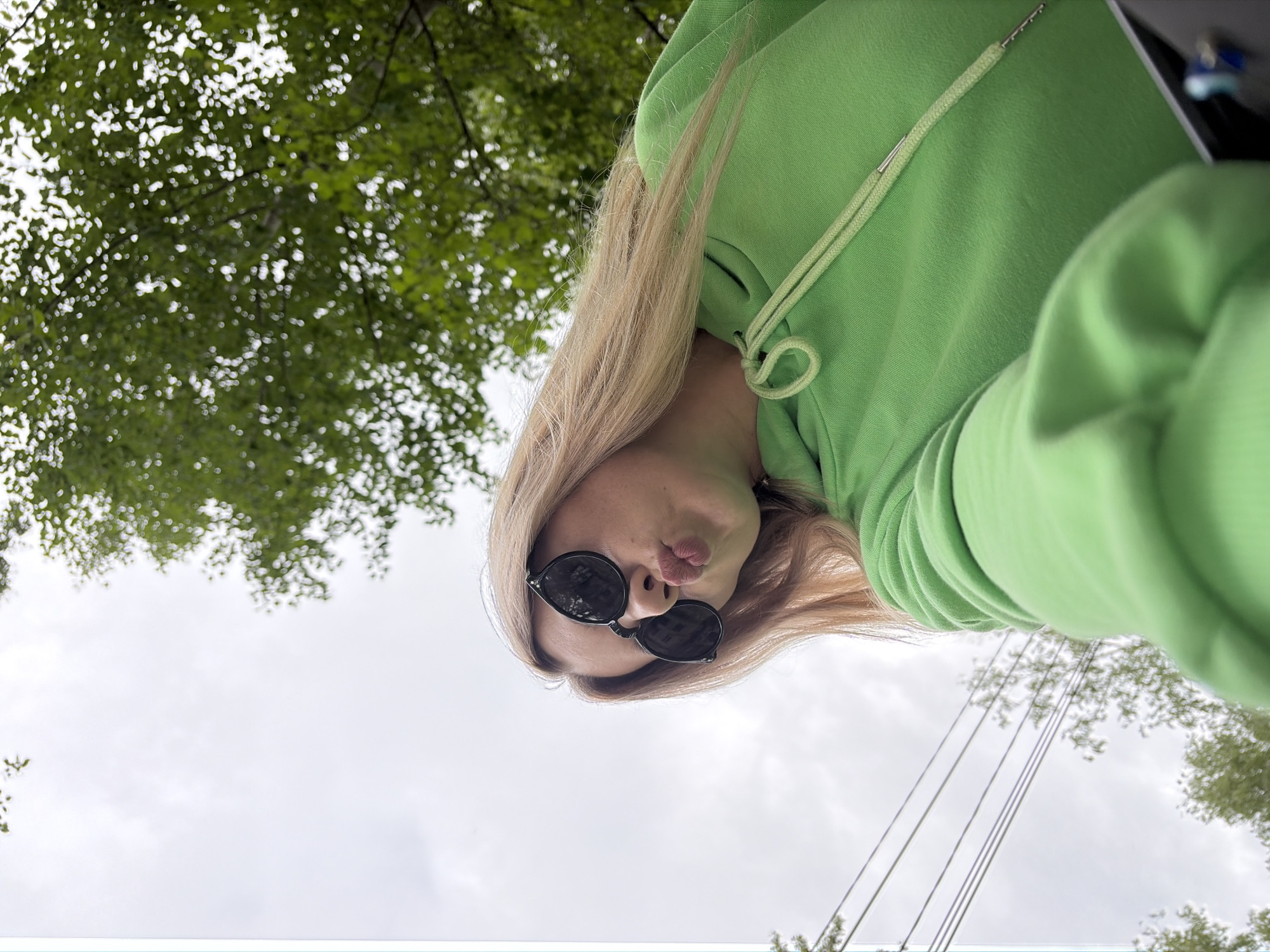 This close-up selfie, captured in Chaikovsky, Russia, features a young woman from a low-angle perspective, looking upwards. She has long, straight blonde hair and is wearing a bright, lime-green hooded sweatshirt with the hood up. Dark, round sunglasses cover her eyes, and her lips are pursed in a playful "kissing" gesture.
The image is rotated, positioning the woman's head towards the upper right, while her body extends diagonally towards the lower left. The background consists predominantly of a bright, overcast sky. Patches of green tree foliage are visible in the upper left corner and sparsely in the lower right, indicating an outdoor setting. Several thin, dark wires, possibly utility lines, traverse the sky in the lower right portion of the frame. The scene takes place during the daytime under what appears to be cloudy weather conditions. No text is visible in the image.
