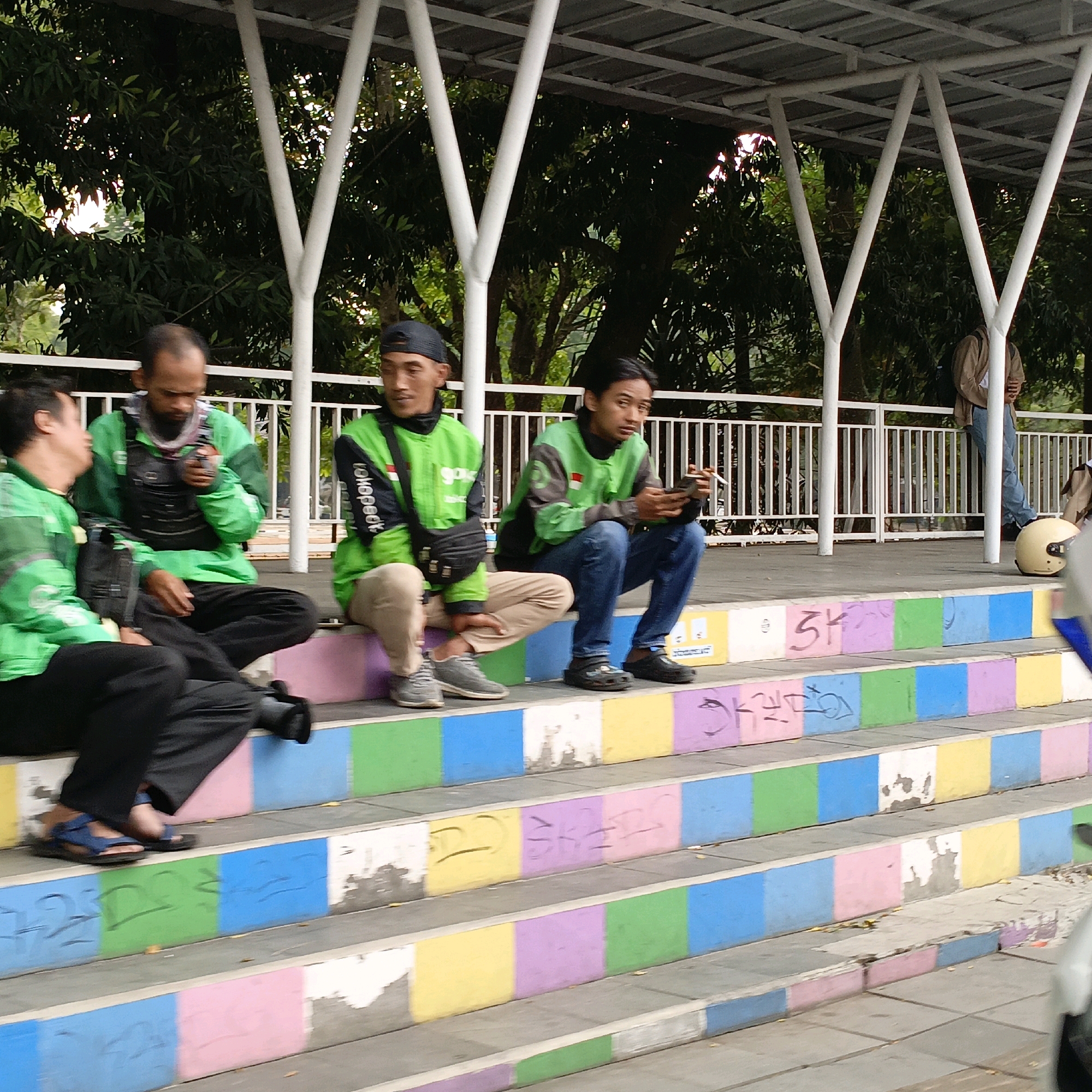 The image depicts a group of five men, presumably motorcycle taxi drivers, resting on a set of multi-colored, concrete steps in Salatiga, Indonesia. The men are dressed in green jackets, commonly worn by ride-sharing drivers in the region.
The man on the far left, wearing black pants and blue sandals, is seated with his legs extended. Next to him, a man in a green jacket and black vest is looking down, holding a smartphone in his left hand and possibly a cigarette in his right. To his right, another man in a green jacket and black pants sits with his legs bent, looking towards the right. The fourth man from the left, also in a green jacket and beige pants, is seated with his hands in his lap, looking down. The man on the far right, in a green jacket and blue jeans, is crouched on the steps, holding a smartphone and a cigarette.
The environment suggests an outdoor public space. Behind the men is a white metal railing and dense foliage, indicating trees or other vegetation. Above them, a white structure with a metal roof provides shade. The steps themselves are painted in alternating blocks of color: pink, blue, green, yellow, and white. Some of the painted surfaces show signs of wear and tear, with graffiti visible on some of the colored sections. A beige motorcycle helmet is placed on the ground to the right of the steps. The overall impression is a casual gathering of workers taking a break in an urban setting.