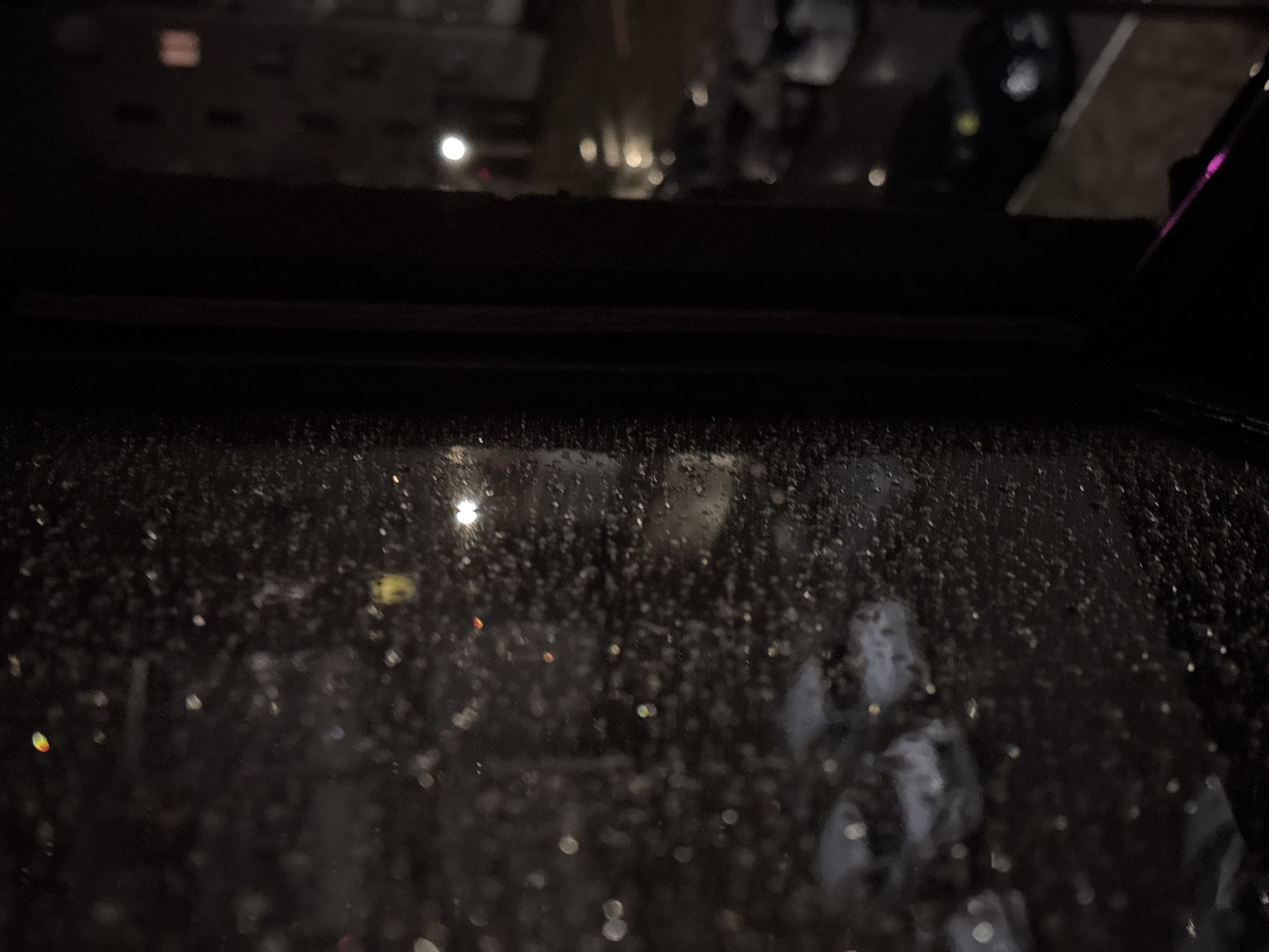 The image depicts a dark, nocturnal scene viewed through a window heavily covered in water droplets or condensation. A thick, dark horizontal frame spans the upper portion, suggesting an interior vantage point, possibly from a vehicle or building.
Beyond the wet glass, the background is deeply out of focus, rendering exterior elements as soft, blurred light sources. Numerous artificial lights, appearing as bokeh, are visible, predominantly white and yellow, with hints of red, orange, and magenta from specific light sources. These scattered lights suggest an urban or populated environment. A particularly bright, star-like light is prominent in the upper-right section of the view through the glass.
In the lower-right area of the wet window, faint, light grey, abstract shapes are discernible. These forms are highly distorted by the water and blur, presenting a somewhat ethereal or ghostly appearance, possibly reflections of interior objects or very indistinct exterior elements. The overall ambiance is one of observing a rainy, illuminated night in Марковский, Russia, from a sheltered position. No text is visible in the image.
