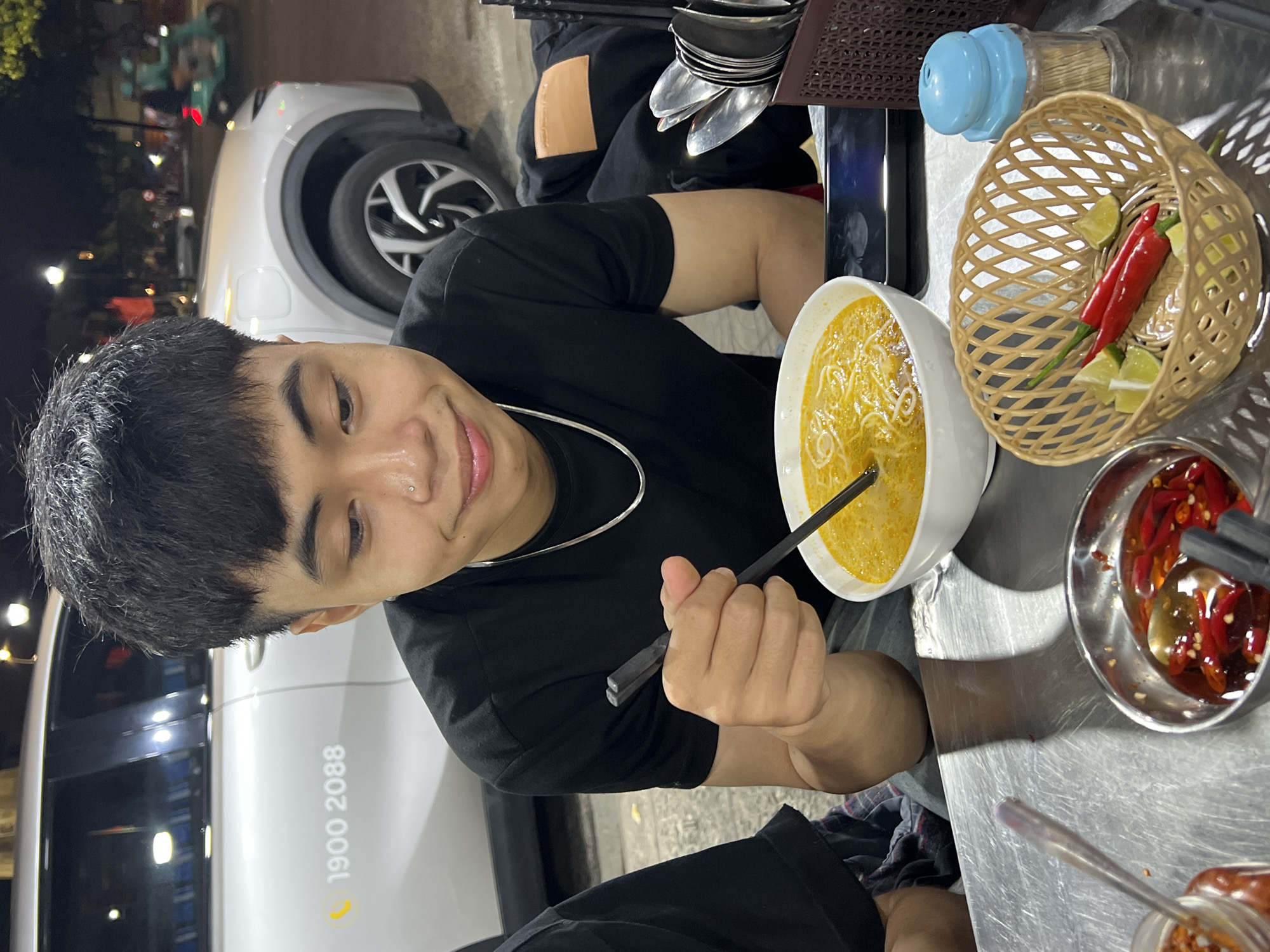 A person sits at a metal table outside at night. The person is holding a pair of black chopsticks in their right hand and stirring a bowl of yellow noodle soup. The person is wearing a black t-shirt and a silver chain necklace. On the table are a bowl of chili peppers, a basket of limes and red chilies, a container of toothpicks, and a stack of spoons and forks. A white car is parked behind the person, with the phone number 1900 2088 visible on the side. The scene is likely in Đà Nẵng, Vietnam, based on the style of food and the phone number.