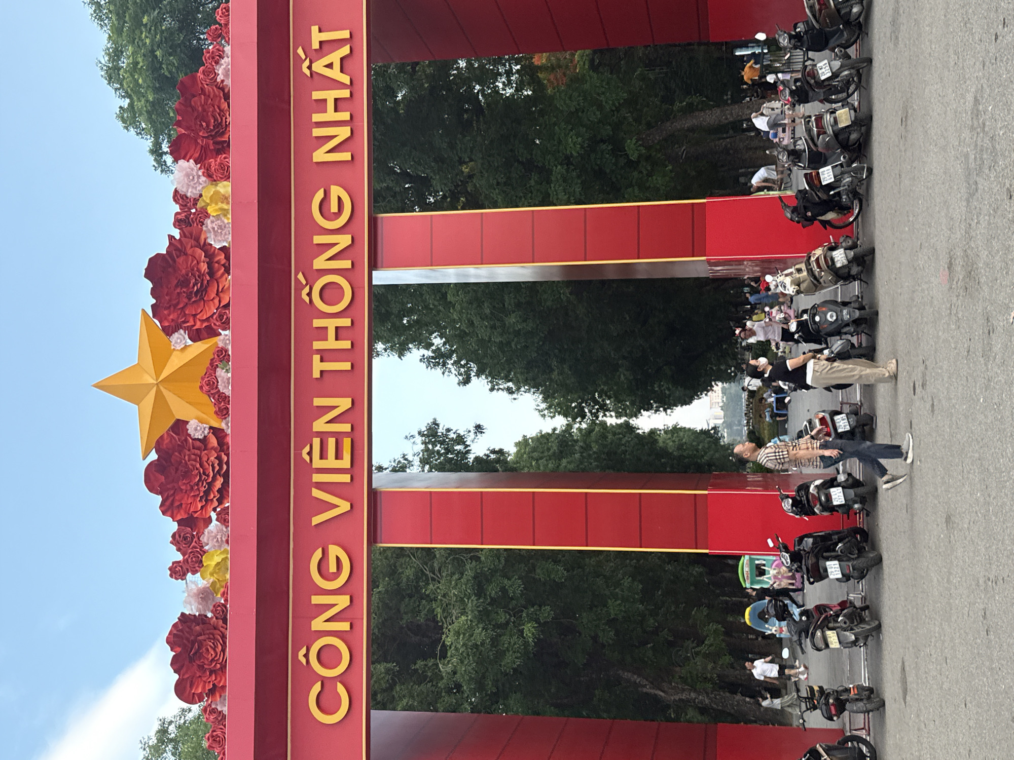 Here is a detailed description of the image:
The image shows the entrance to Thống Nhất Park (Công viên Thống Nhất) in Hà Nội, Vietnam. A large red archway with yellow lettering spells out the park's name, decorated with red and other colorful flowers and a yellow star. Rows of motorbikes are parked along the side of the road, and people are walking around. The background features lush green trees. The scene appears to be a typical day at the entrance of a popular urban park.