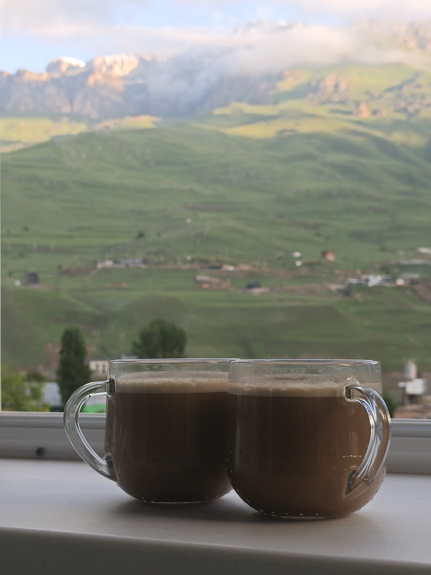 Two clear glass mugs filled with a frothy, light brown liquid, likely coffee or a similar beverage, sit on a white windowsill. The mugs have curved handles. In the background, a hazy, green mountainous landscape is visible under a sky with scattered clouds. The slopes of the mountains are dotted with small buildings and patches of trees, suggesting a rural or semi-rural setting. The lighting suggests it may be early morning or late afternoon. The scene evokes a peaceful, serene atmosphere. The location is Verkhny Fiagdon, Russia.