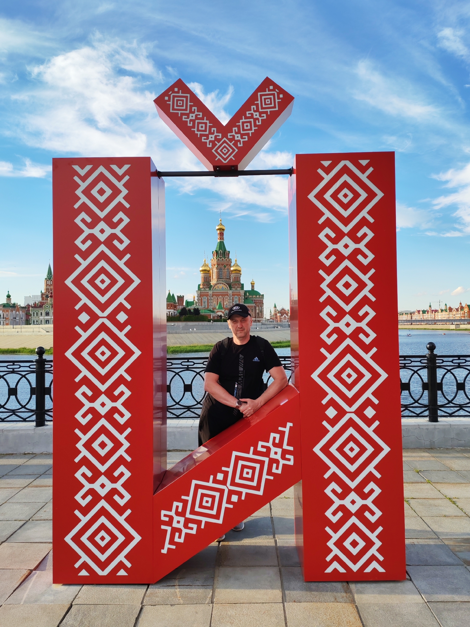A man stands in front of a large red letter "Н" decorated with white traditional patterns. The letter is a prominent object in the scene, forming a frame for the man and the background. The man is wearing a black t-shirt, black shorts, and a black baseball cap. He is leaning on the lower part of the letter "Н" with his arms crossed.
The background features a scenic view of a city along a river. Several ornate buildings with golden domes and colorful facades are visible across the water, including what appears to be a cathedral. A bridge spans the river in the distance, and boats are present on the water. A black metal fence with circular patterns lines the embankment in front of the river. The sky above is blue with scattered white clouds, suggesting it is daytime and the weather is fair. The ground in the foreground is paved with gray and beige tiles.
The setting appears to be a public waterfront area, possibly a promenade or park, in Yoshkar-Ola, Russia, given the architectural style of the buildings and the presence of the Cyrillic letter. The overall mood is relaxed and picturesque. Visible text includes "KIA MOTORS" on a building in the background and "ADIDAS" on the man's t-shirt.