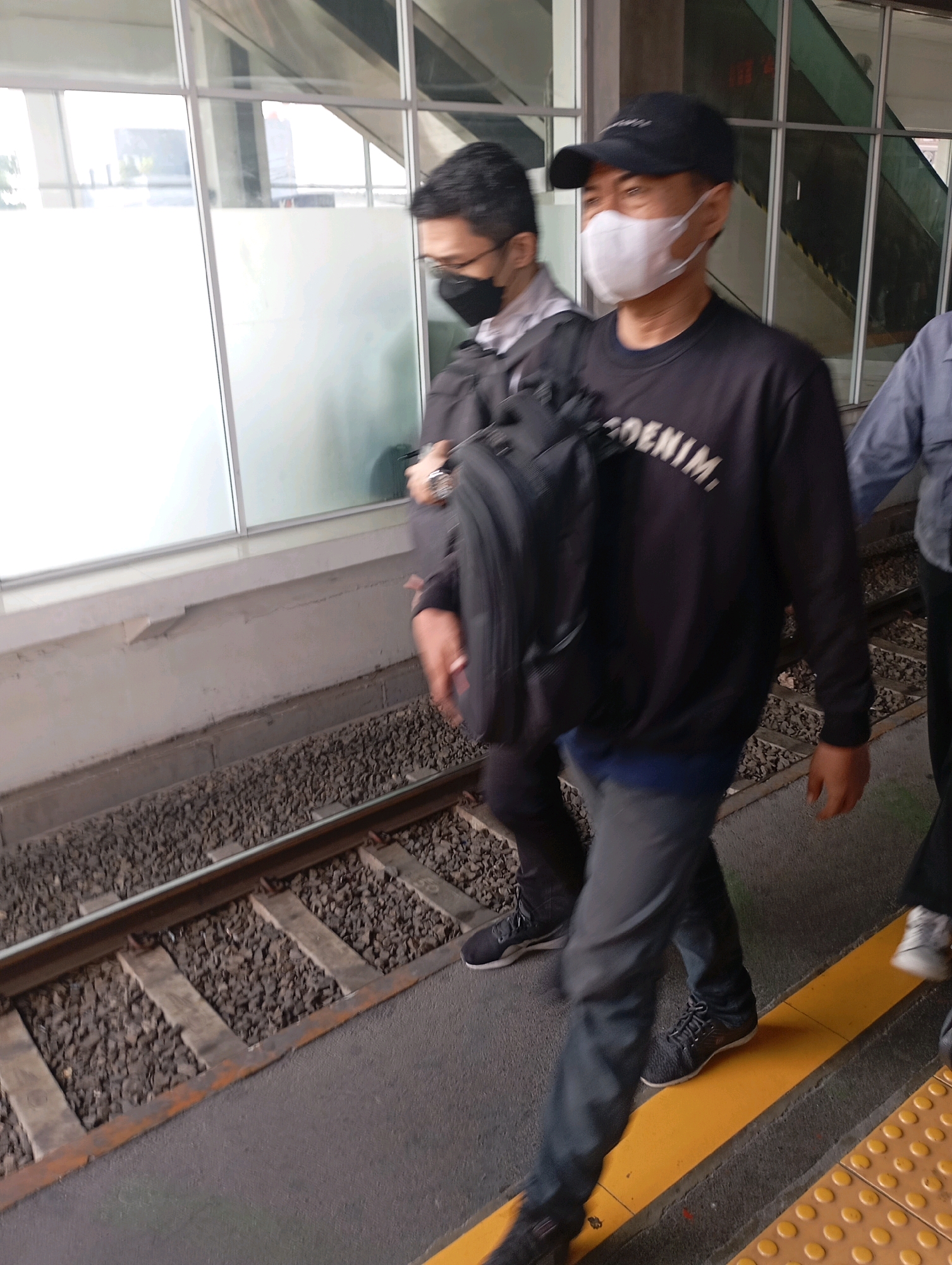 The image depicts a group of people walking along a train platform in West Jakarta, Indonesia. The individuals are wearing masks and casual clothing. One person in the foreground wears a black cap, a dark sweatshirt with "DENIM" printed on it, and grey pants. They are carrying a black backpack. Another person to their left wears a black mask and a light-colored jacket over a dark shirt. The platform features train tracks to the left and a yellow tactile paving strip to the right, indicating it is a public transport area. The environment appears to be an indoor or covered train station with large windows and a muted color palette. The lighting suggests it is daytime.