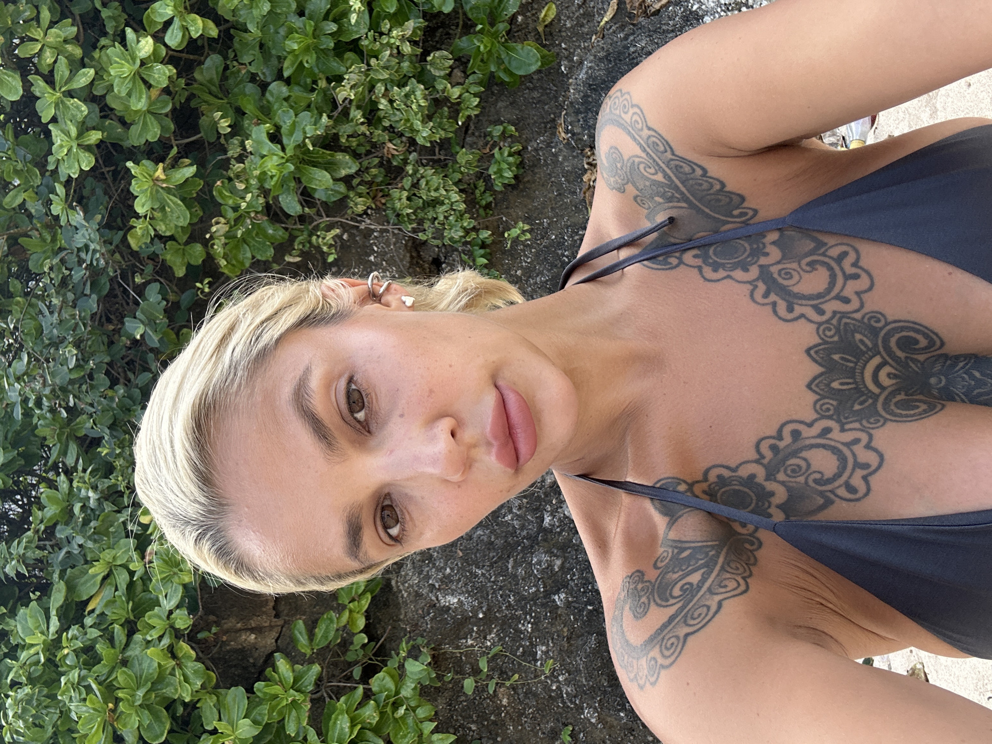 A woman with blonde hair and tattoos is taking a selfie outdoors. She is wearing a dark grey bikini top. She has a heart-shaped earring in her left ear. Behind her is a wall of green foliage and rocks. The scene is likely in Kuta Selatan, Indonesia.