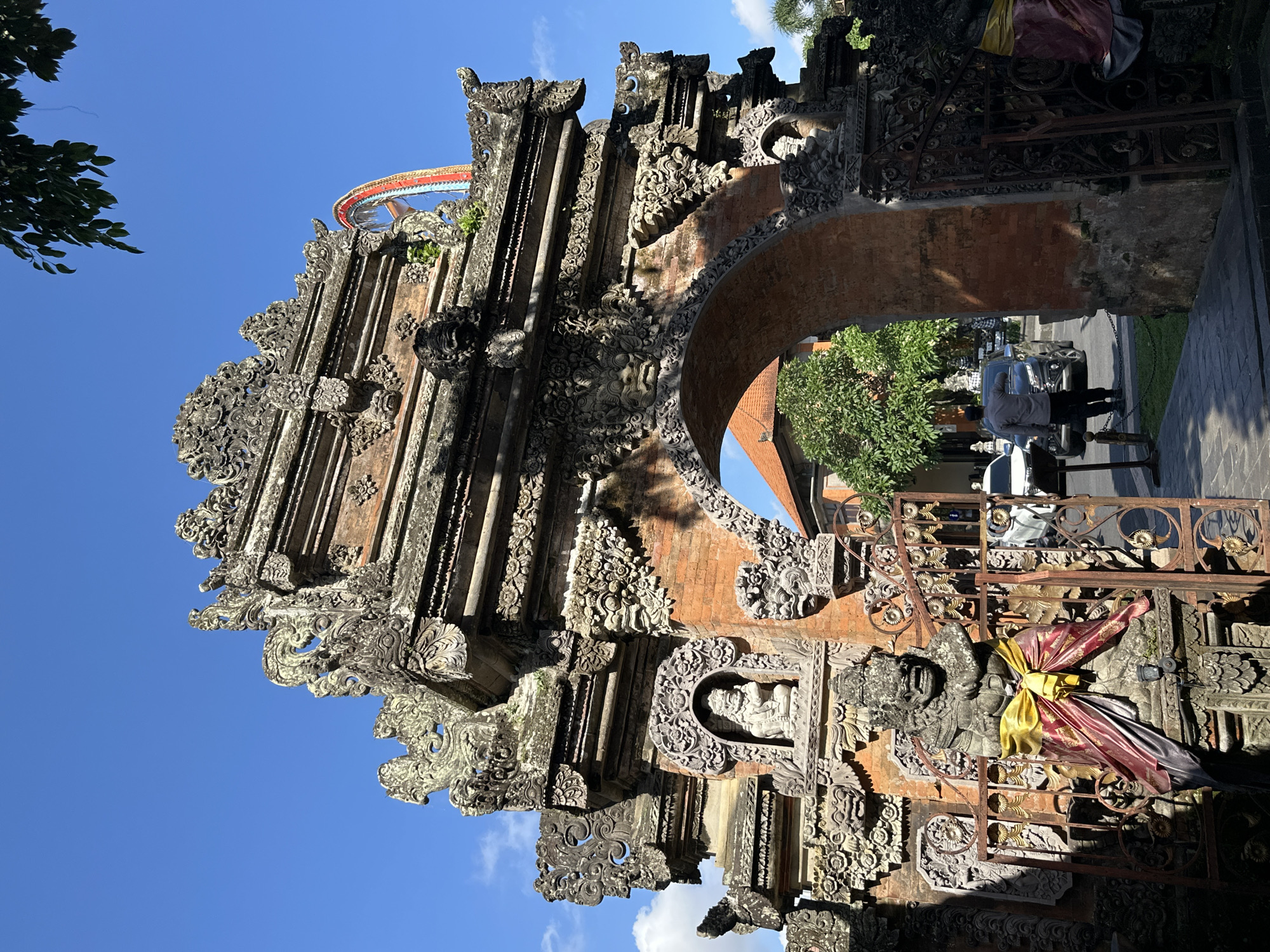 This is an outdoor, eye-level, full-shot photograph of an ornate, traditional Balinese stone archway and gate in Ubud District, Indonesia, under a clear blue sky. The archway, constructed from stone and brick, features intricate carvings of mythical creatures and floral motifs, with moss and weathering visible on its surface. In the foreground, to the left of the gate, stands a stone statue of a fierce demon-like figure adorned with a yellow and red sash. The gate itself is made of dark metal with decorative patterns.
Through the archway, a street scene is visible, with a dark-colored SUV and a white car parked. A man in a gray shirt and dark pants is standing near the dark SUV, facing away from the camera. Trees and the roof of a building are also visible in the background. A segment of a brightly colored, possibly decorative, structure is visible in the upper right quadrant of the image, behind the archway. Sunlight casts shadows on the paved pathway in the foreground, indicating a bright day.