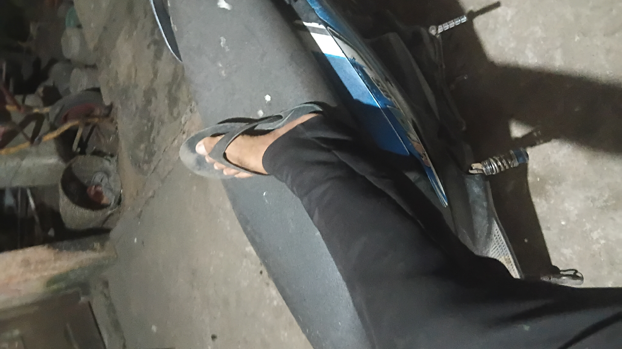 The image shows a person's leg and foot, clad in black pants and a black flip-flop, resting on the seat of a motorcycle. The motorcycle is blue and black, with visible suspension components. The background appears to be an outdoor, unpaved area, possibly a yard or alleyway, with loose gravel or dirt on the ground. In the background, there are several objects that suggest a workshop or storage area: a large, cylindrical object that might be a barrel or a large wheel, some metal structures, and possibly parts of machinery. The lighting suggests it is nighttime or a very dimly lit area, with harsh shadows cast by the objects. The overall impression is of an industrial or utilitarian setting. While the image itself doesn't contain explicit markers of Prabumulih, Indonesia, such settings are common in areas where motorcycles are a primary mode of transportation and where light industry or informal workshops operate.