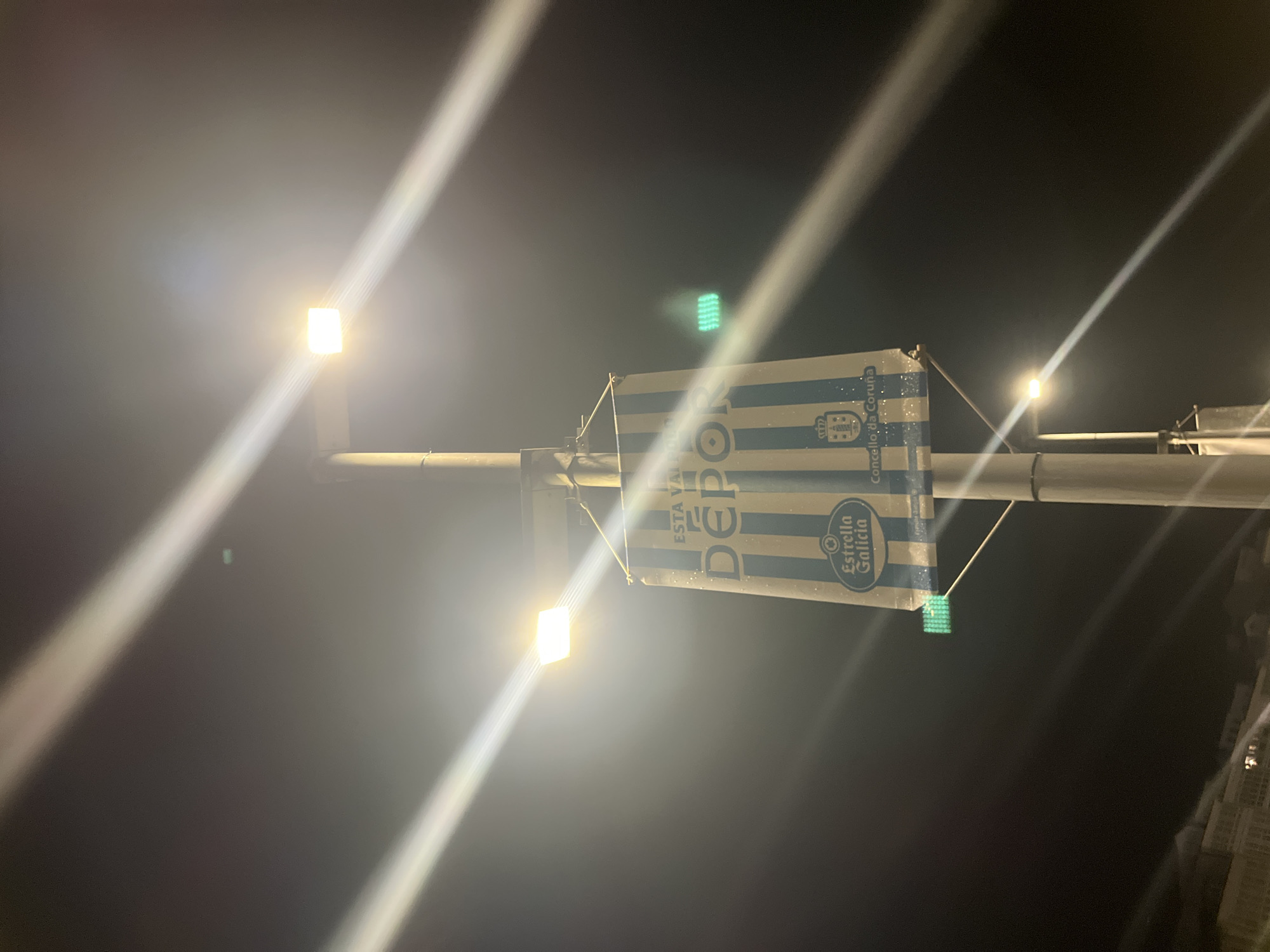 A nighttime image of a street lamp post displaying a flag for the Deportivo La Coruña football club. The flag features the team's blue and white striped colors, the club's crest, the logo of Estrella Galicia (a local beer brand), and the inscription "ESTA VALE DEPOR" (a club slogan). The flag also has the emblem of Concello da Coruña (City Council of A Coruña). The image is taken at night, with the street lamps providing the primary source of illumination.