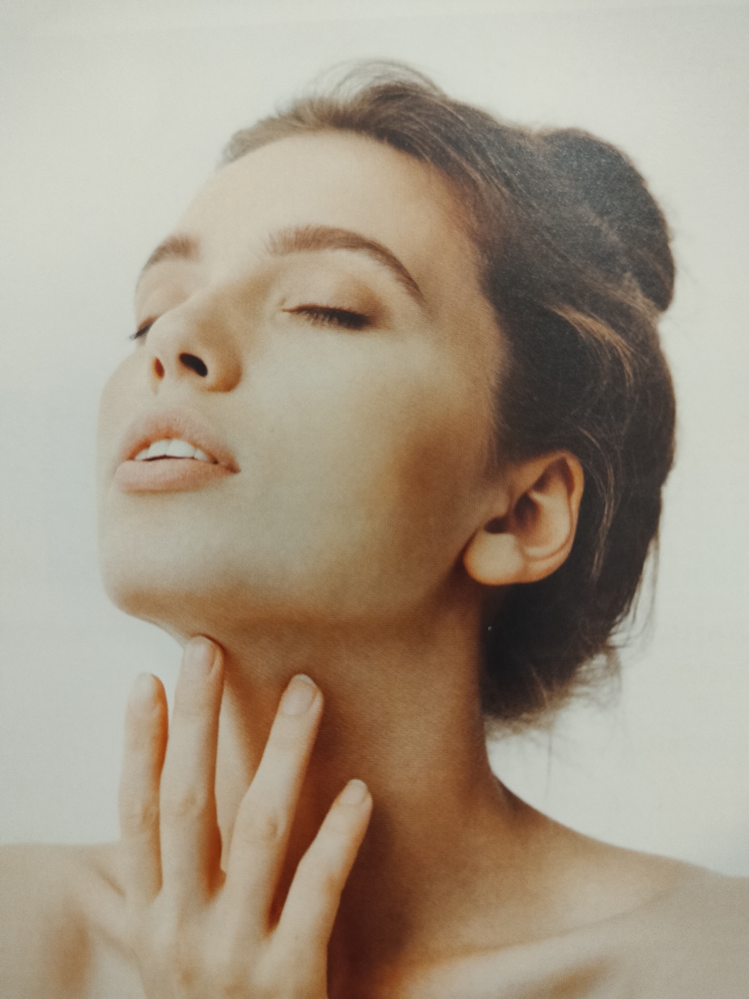 The image is a close-up portrait of a young woman with her eyes closed and her head tilted back. Her right hand is gently touching her neck and chin. The background is a plain, light color, suggesting a studio setting. There is no visible location in Novosibirsk, Russia.