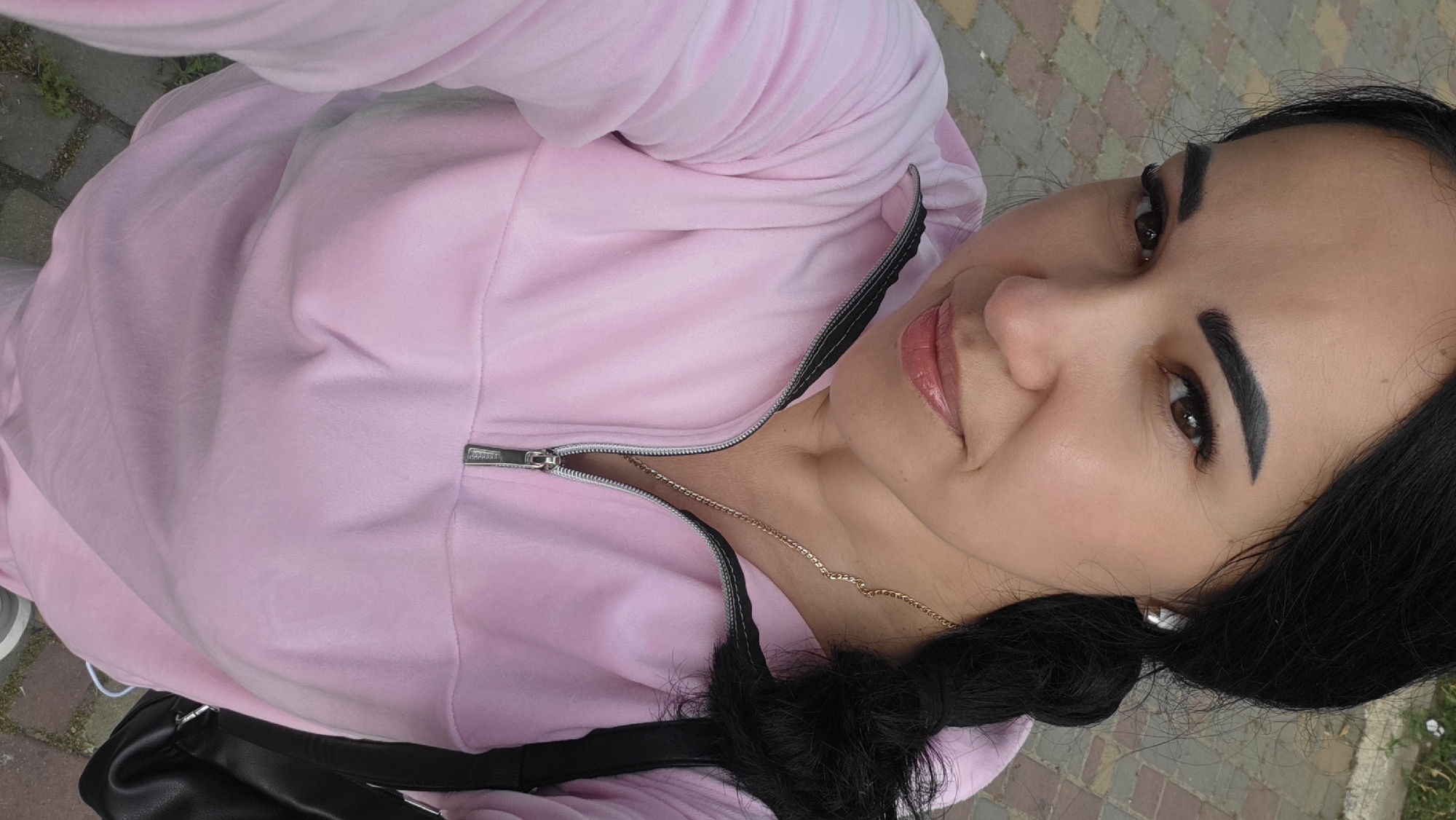 A close-up selfie of a woman in a pink velour hoodie. She has dark hair styled in a braid and is wearing a gold chain necklace and a white earbud. The background appears to be an outdoor paved area, possibly a sidewalk or park.