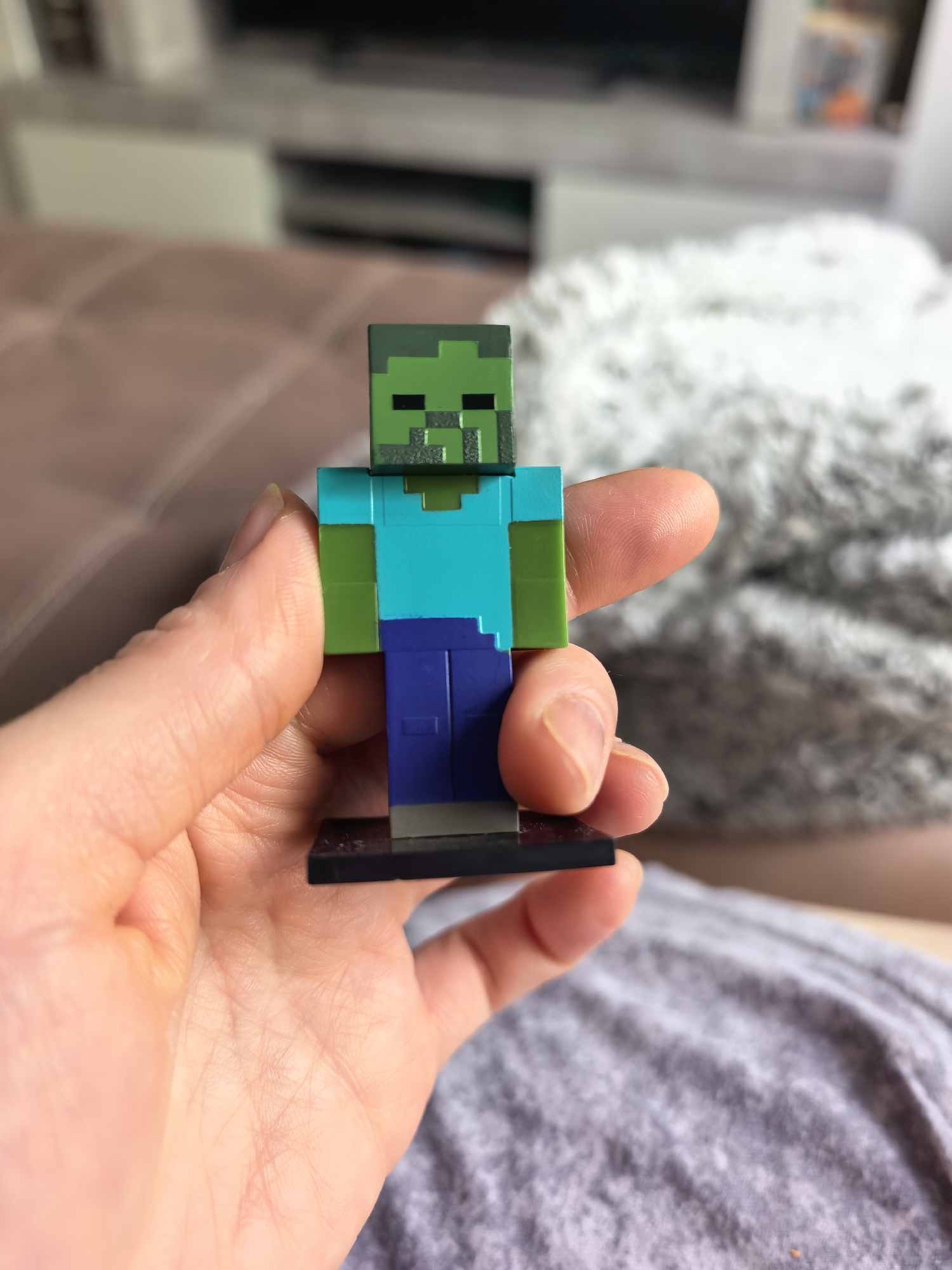 A person is holding a Minecraft zombie figurine. The figurine is green and blue and is standing on a black base. The person is sitting on a gray surface, and there is a couch and television in the background. There is no evidence in the image to suggest the location is in Sterlitamak, Russia.