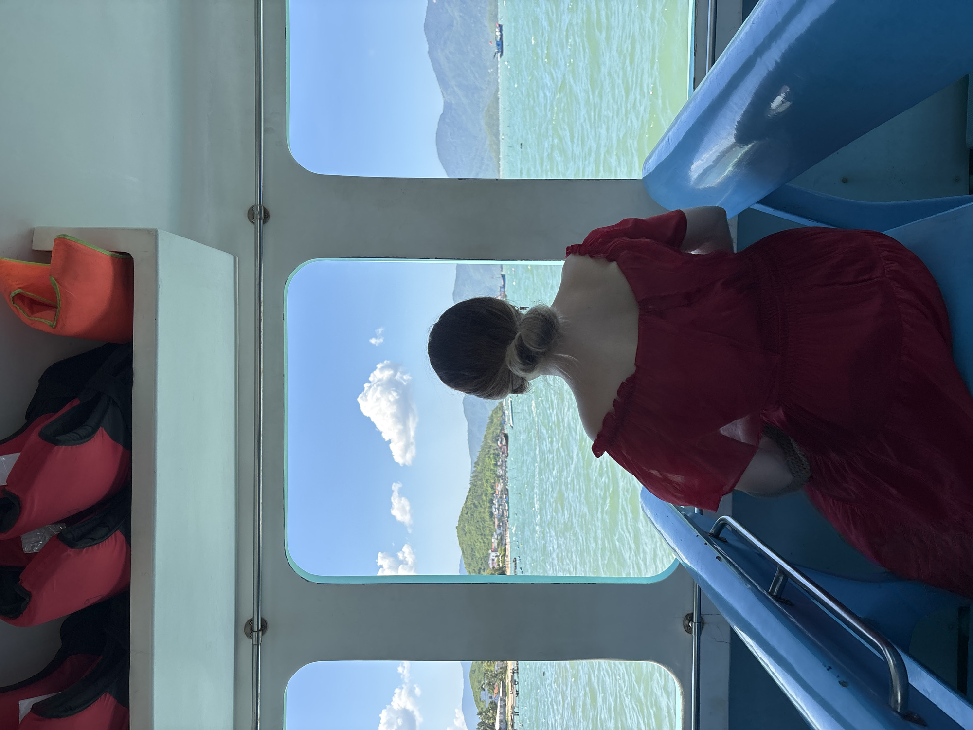 A woman, seen from the back, sits on a blue seat inside a boat, facing three large windows. She is wearing a flowing red dress with short, puffed sleeves. Her hair is styled in a bun. Through the windows, a bright blue sky with scattered white clouds is visible. The ocean, a vibrant turquoise, stretches out to a coastline with green, mountainous islands dotted with buildings and small boats. The interior of the boat has a light blue trim around the windows and blue seating. Red and orange life vests are stored on a shelf above the windows. The scene appears to be a scenic view from a boat traveling near Bac Nha Trang Ward, Vietnam.