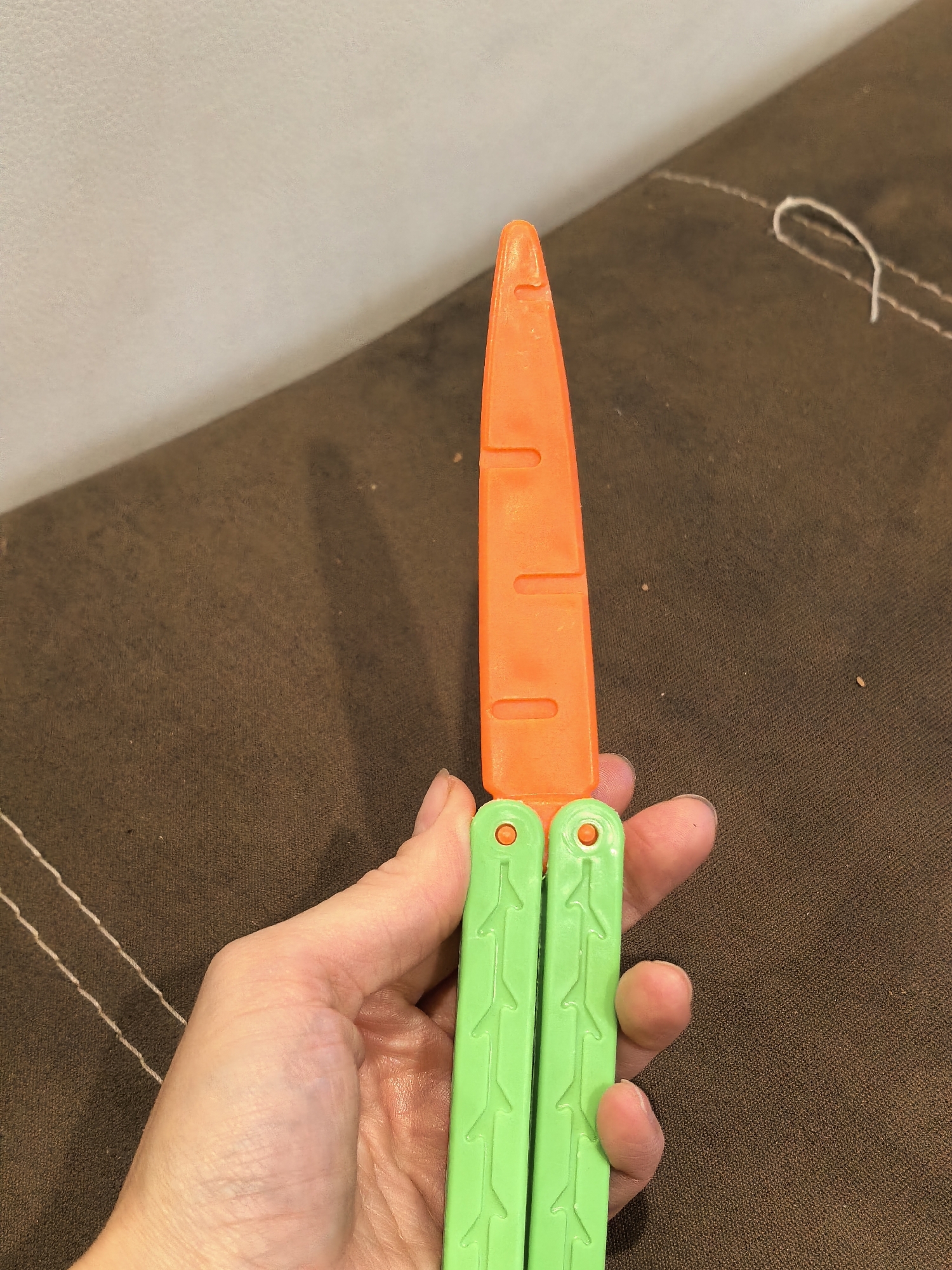 A person's hand is holding a brightly colored toy that resembles a carrot on a butterfly knife handle. The toy carrot is orange with cut marks to simulate segments, and the handle is bright green with textured details and circular protrusions. The background is a dark brown, textured surface, possibly upholstery, with some white stitching visible. The lighting suggests an indoor environment. No specific location in Sterlitamak, Russia can be determined from the image.