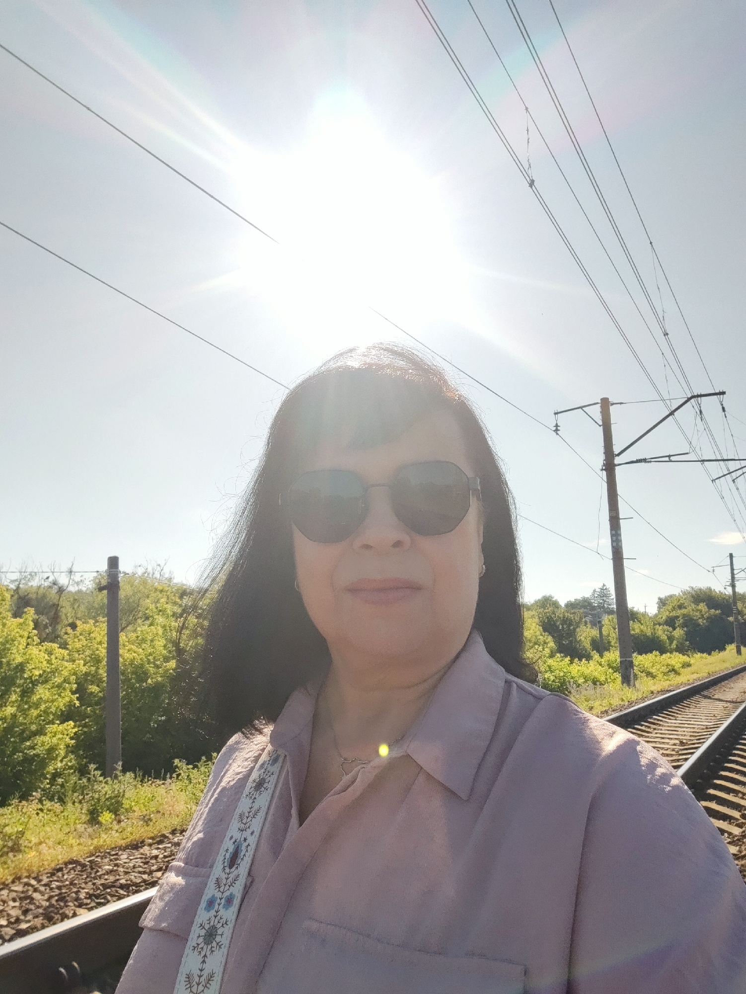 A middle-aged woman with dark hair and sunglasses is taking a selfie outdoors. She is wearing a light purple collared shirt and a light-colored bag with a floral pattern is slung over her shoulder. She is standing next to railroad tracks, with lush green trees and vegetation in the background. Overhead, power lines are visible against a bright, sunny sky, with the sun creating a lens flare. The image appears to be taken on a clear, warm day.