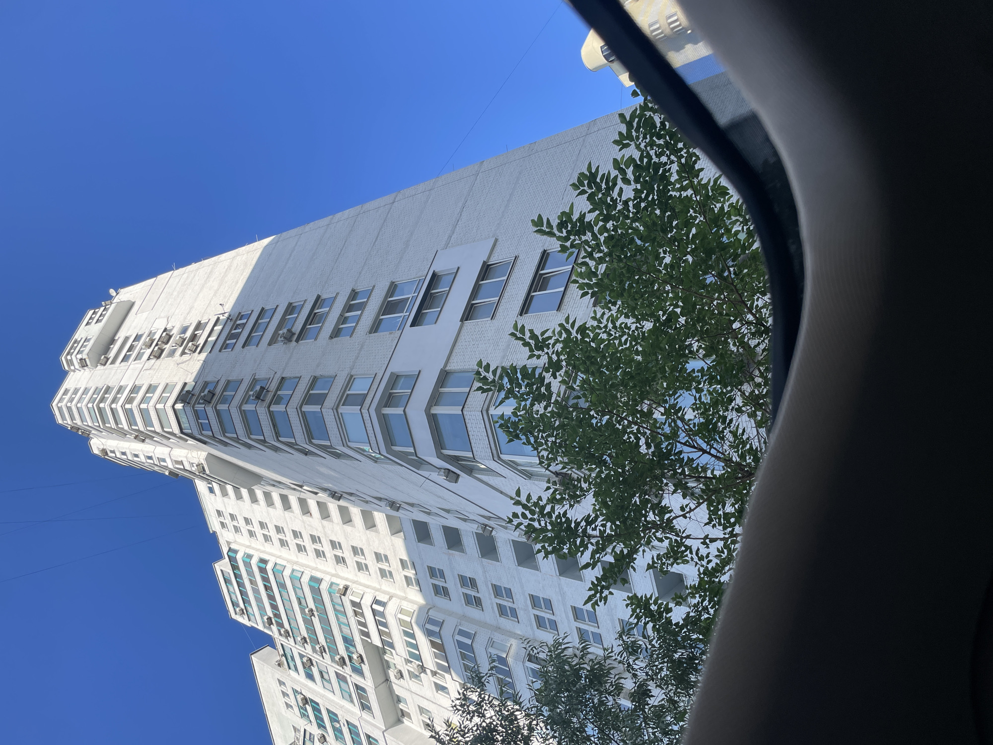 This is a low-angle, exterior, eye-level shot of a modern, white, high-rise apartment building in Kyiv, Ukraine, under a clear blue sky. The building features numerous windows and balconies, many equipped with air conditioning units. Green trees with dense foliage partially obscure the lower sections of the building. In the foreground, a dark, curved object, likely the interior of a vehicle's sunroof or frame, occupies the bottom portion of the image, framing the view of the building and sky. The lighting suggests it is daytime, with sunlight casting shadows on the building's facade.
