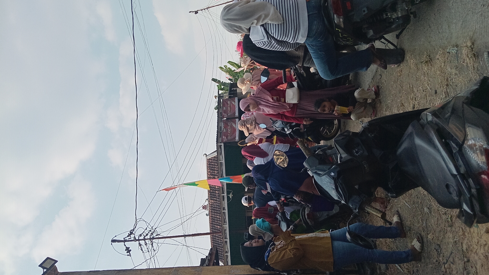 The image depicts a gathering of people in an outdoor setting, possibly a street or open area, in Kedungwaru, Indonesia. The scene is crowded with individuals, many of whom are women wearing hijabs and modest clothing. Several motorcycles are parked and visible, suggesting transportation or a local event. In the background, there are buildings with corrugated roofs and banners displaying text, possibly advertisements or event information. Utility poles with numerous wires are also prominent in the sky. The ground appears to be dirt and gravel, with some scattered dry grass or straw. A young girl is seen squatting and drinking from a cup. The lighting suggests it is daytime, with a partly cloudy sky.