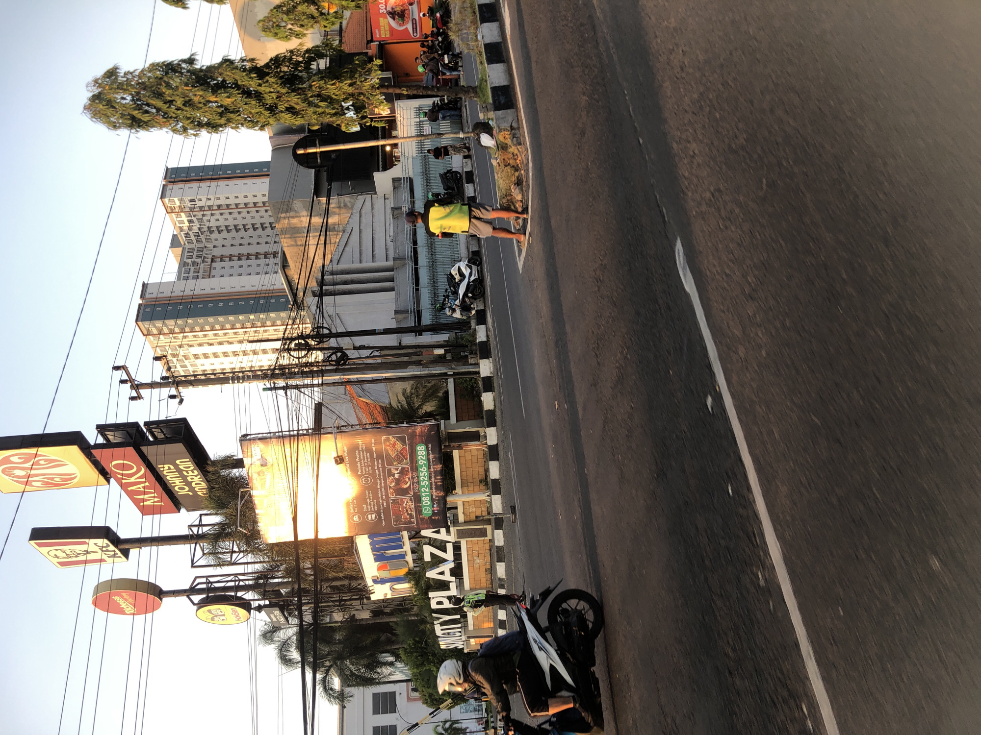 The image depicts a street scene in Sidoarjo, Indonesia, likely near Sun City Plaza. In the foreground, a person rides a white motorcycle to the left, wearing a dark jacket and a white helmet. To the right, on the sidewalk, a person in a bright yellow and black vest stands facing away from the camera, wearing gray shorts and flip-flops. Behind them, a few other individuals are gathered, some near motorcycles.
Dominating the background is a tall, multi-story building with a light-colored facade and numerous windows. Several tall trees are also visible, including one prominent, slender tree on the right. Various signs are present, including a prominent KFC sign, a red circular sign with "Richeuse," a sign for "MAKO JOHNNY ANDREAN," and a large billboard for "Sun City Plaza" which includes promotional information and a phone number. Electrical wires crisscross the scene, indicating urban infrastructure. The overall impression is a busy street during daylight hours, likely late afternoon due to the warm lighting.