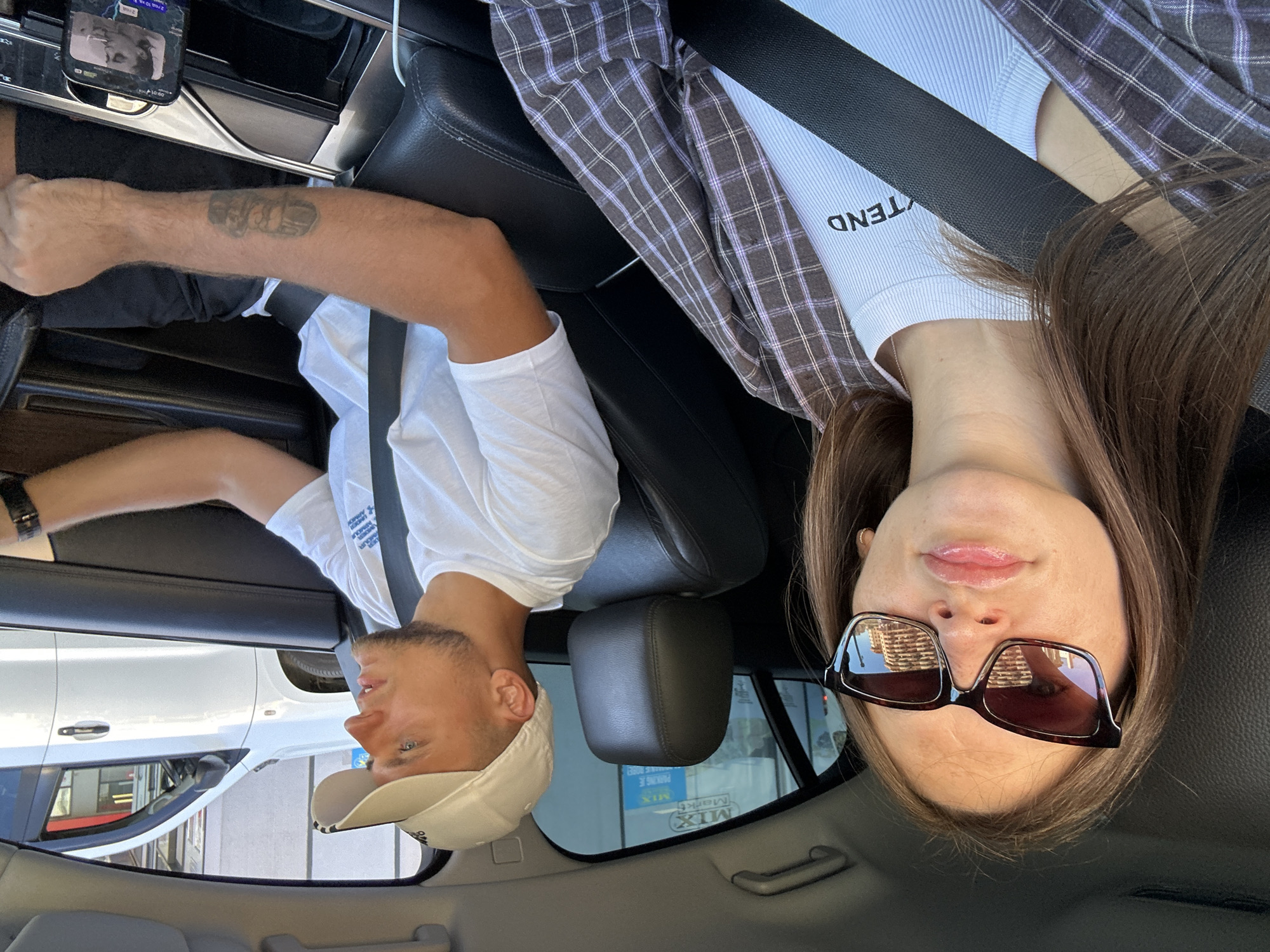 In Donji Pobori, Montenegro, a man wearing a white baseball cap and a white t-shirt is driving a car. He has a tattoo on his right forearm. Next to him, a woman wearing sunglasses and a plaid shirt is looking towards the camera. She is wearing a white ribbed top with the word "TEND" printed on it. The car's interior, including black leather seats and seatbelts, is visible. Outside the car windows, buildings and other vehicles can be seen. A smartphone is mounted on the dashboard, displaying a video of a woman.