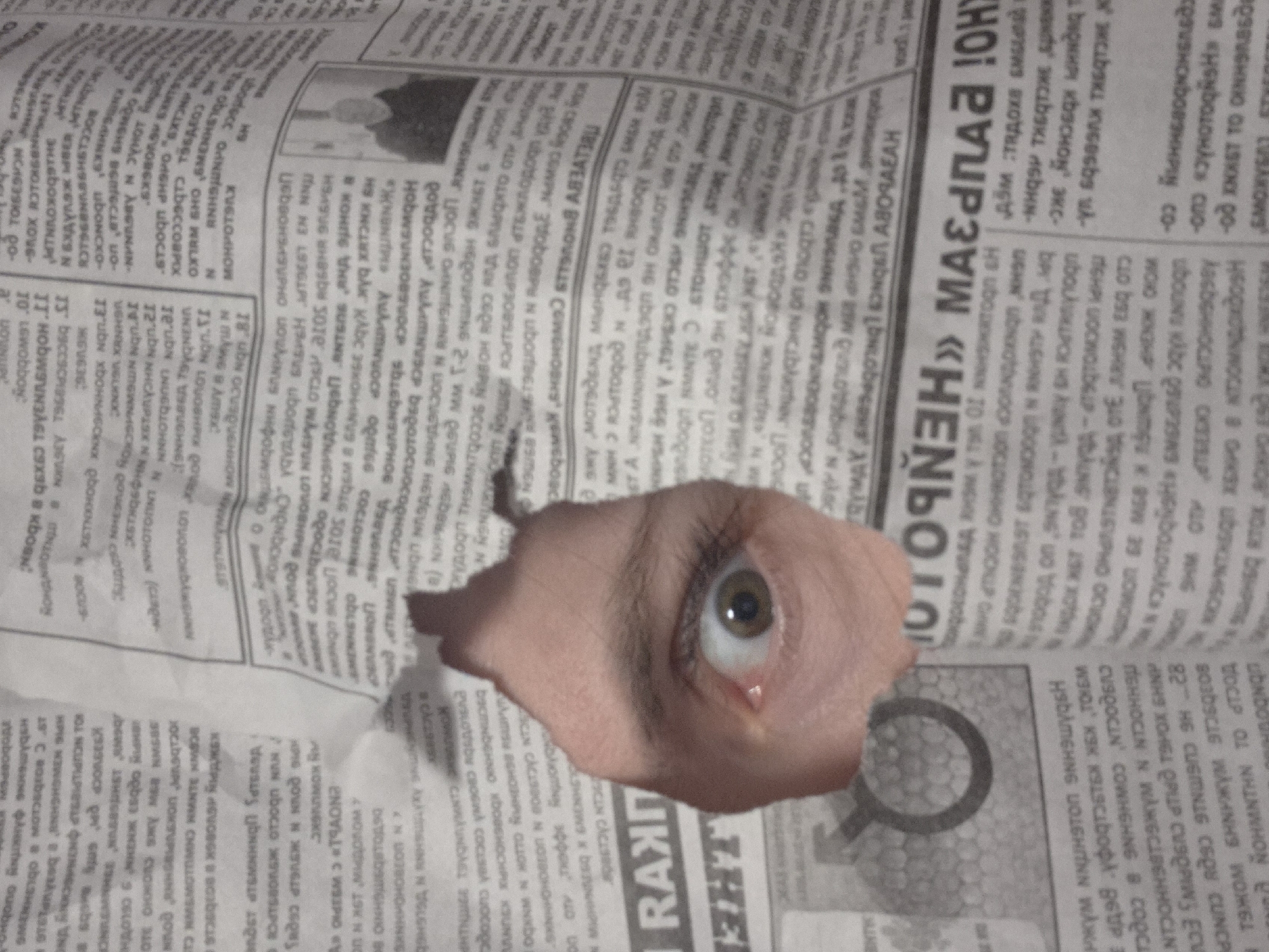 An eye peers through a hole torn in a newspaper, revealing flesh-toned skin around the eye. The newspaper text is in an unidentified language.