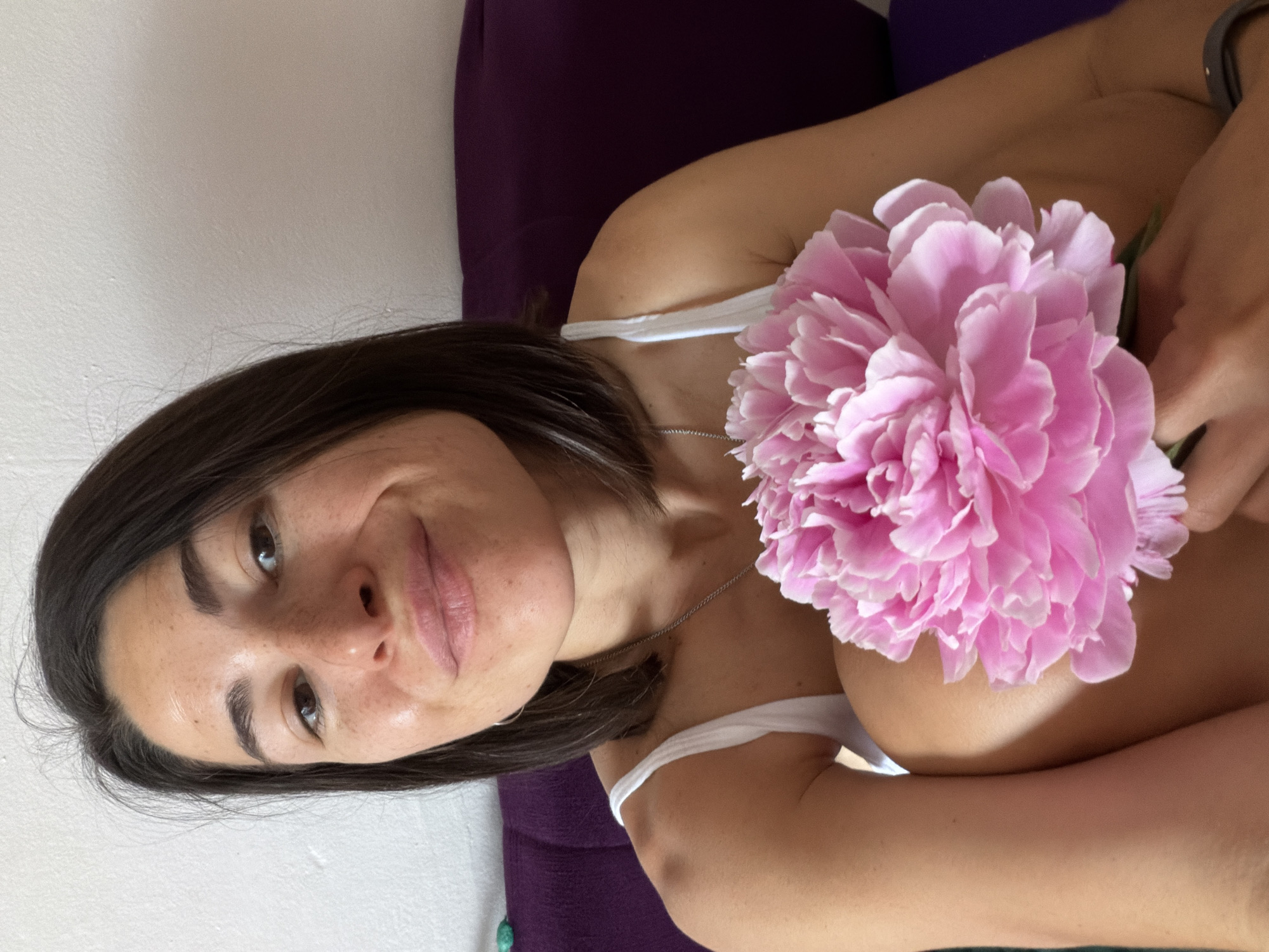 A woman with dark hair and freckles is holding a large pink flower in front of her chest. She is wearing a white tank top and a silver necklace. She is sitting on a purple cushion. The background is a white wall. There is no evidence to suggest this image was taken in Budva, Montenegro.