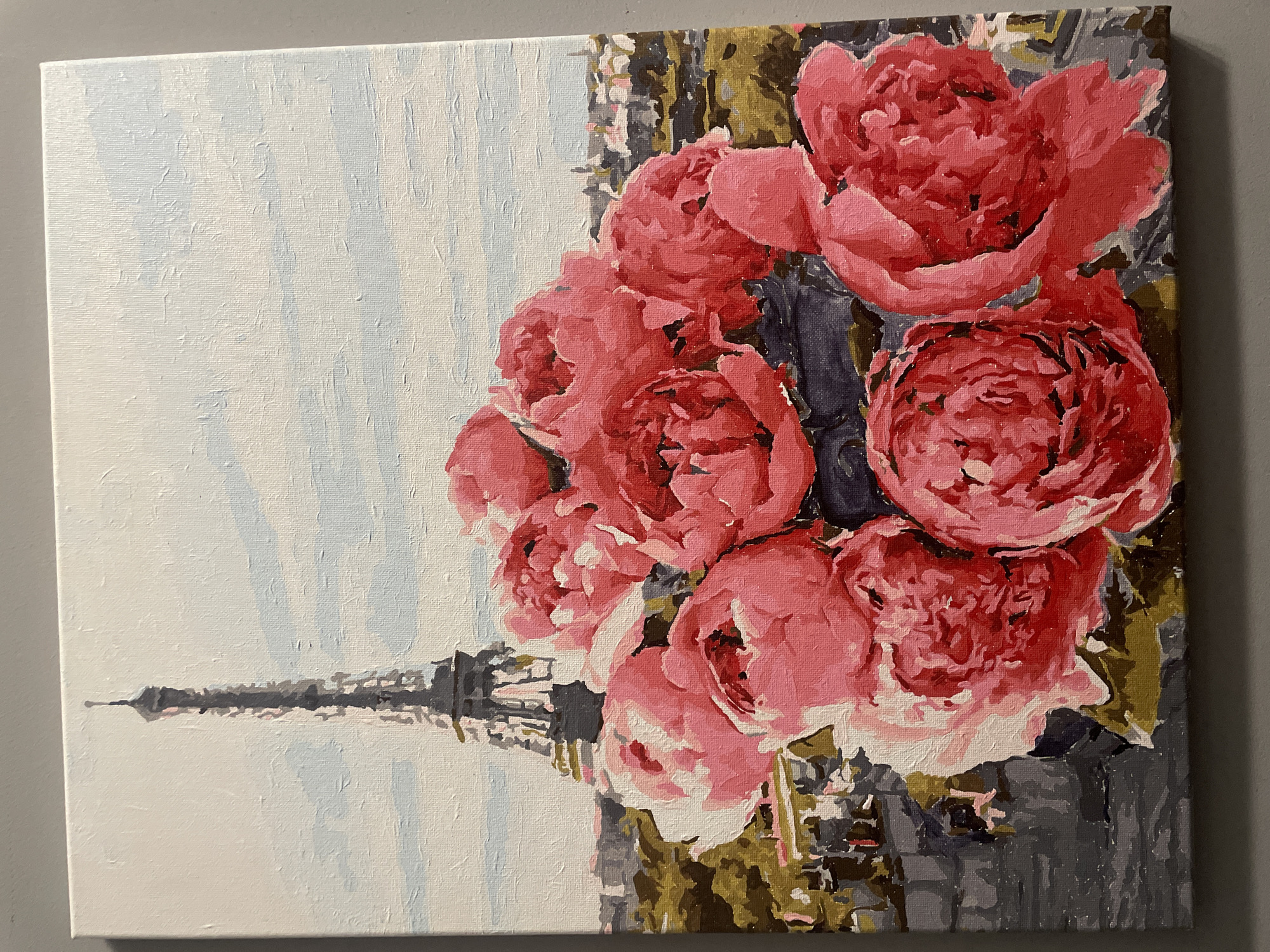 The media file displays a painting on canvas, likely acrylic, featuring a stylized composition. The painting exhibits characteristics of a "paint by number" or similar art style, with distinct blocks of color and visible brushstrokes, particularly evident in the close-up crops.
The primary subjects are a large, vibrant bouquet of pink and red flowers, resembling peonies or roses, which dominate the right side of the canvas. These flowers are rendered with various shades of pink and red, creating a textured and somewhat abstract appearance.
On the lower-left side of the canvas, the recognizable silhouette of the Eiffel Tower is depicted against a lighter, possibly sky-blue and white background. The tower is painted in shades of gray and black, suggesting it is further in the background relative to the prominent flowers. The background directly behind the flowers on the right is composed of muted greens, grays, and dark blues, appearing as abstract foliage or urban structures.
The painting's composition balances the large, foreground floral elements with the more distant architectural landmark. The overall impression is one of a romanticized scene. There is no explicit activity taking place; it is a still life or landscape. No visible text is present in the image.
The prominent inclusion of the Eiffel Tower strongly suggests the location depicted is Paris, France. This contradicts the provided location context of Sochi, Russia.