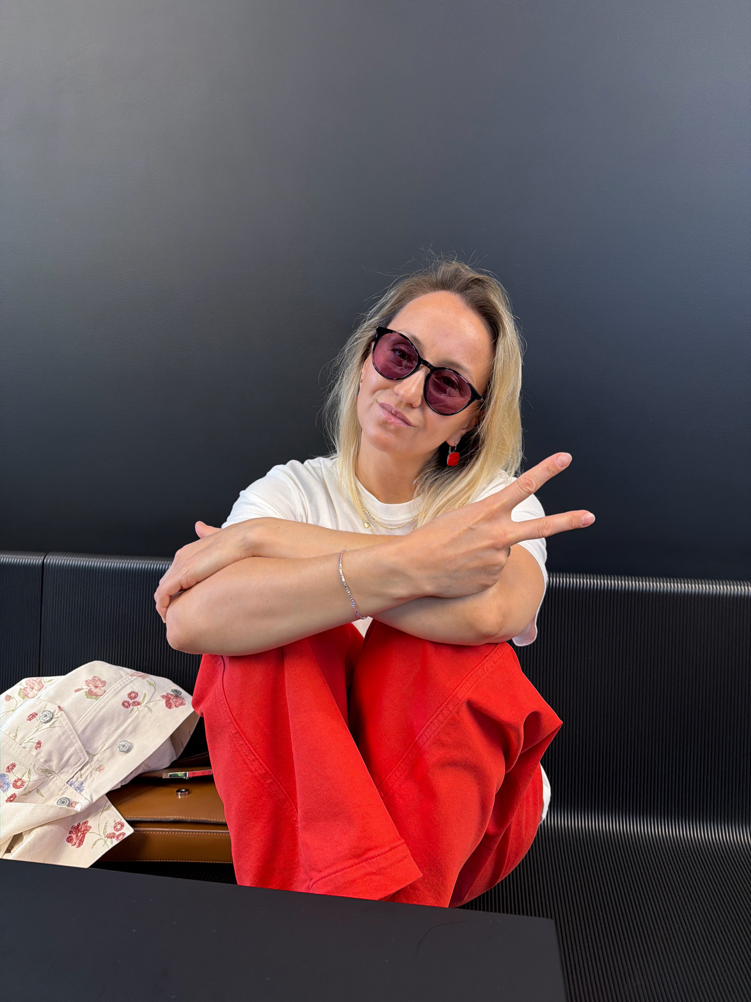 A Caucasian woman with blonde hair and sunglasses is sitting on a black bench. She is wearing a white t-shirt, red pants, and red earrings. Her arms are crossed, and she is holding up two fingers in a peace sign. To her left is a patterned jacket and a brown briefcase. The background is a dark gray wall. The image was taken in Izhevsk, Russia.