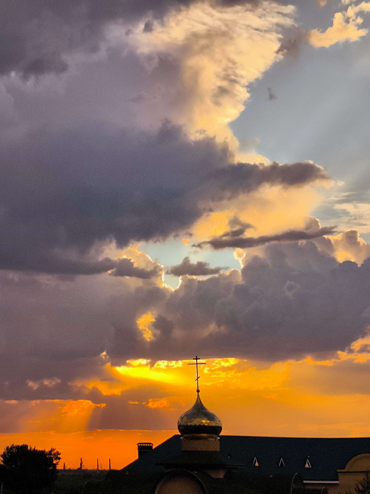 The image shows a dramatic sunset over a city, likely Qarağandı, Kazakhstan. The sky is filled with vibrant orange and yellow hues from the setting sun, peeking through dramatic, dark clouds. Beams of light are visible piercing through the cloud cover. In the foreground, the golden dome and cross of an Orthodox church are silhouetted against the bright sky. The roof of another building, possibly part of the same complex, is visible below the dome. In the distance, a dark silhouette of trees and industrial structures can be seen against the horizon. The overall scene conveys a sense of awe and natural beauty.