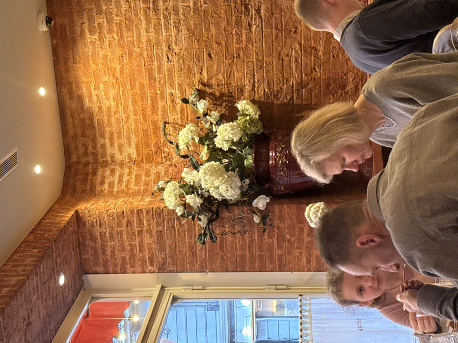The scene depicts the interior of a dining establishment, identified as being in Paris, France. The number "164" is visible on a building across the street, reflected through a large window on the left side of the room, along with the inverted text "RICHE" on the glass.
The interior features reddish-brown brick-patterned walls and a white ceiling equipped with multiple recessed spotlights and a linear air vent. A white dome-shaped security camera is mounted on the ceiling in the upper right area. Centrally positioned against the brick wall is a large, ornate dark red vase containing an arrangement of artificial white and light green flowers, resembling hydrangeas, along with dark green foliage. A red fabric valance or awning is visible above the top of the window on the left.
Four individuals are partially visible, seated at a table in the foreground. In the immediate foreground-left, a man with short, dark hair, wearing a light brown or grey collared shirt, is seen from his back and right side, holding a spoon to his mouth, suggesting he is eating. To his left and slightly behind him, a young girl with light brown, curly hair, wearing a light pink top, looks towards the left, possibly holding cutlery. In the middle-right of the frame, a woman with shoulder-length blonde hair, wearing a grey sweater and a silver necklace, is seen in profile, looking downwards. To her right, the back of a second man's head and shoulder are visible; he has short, light brown hair and is wearing a dark blue or black garment, also looking downwards or straight ahead.