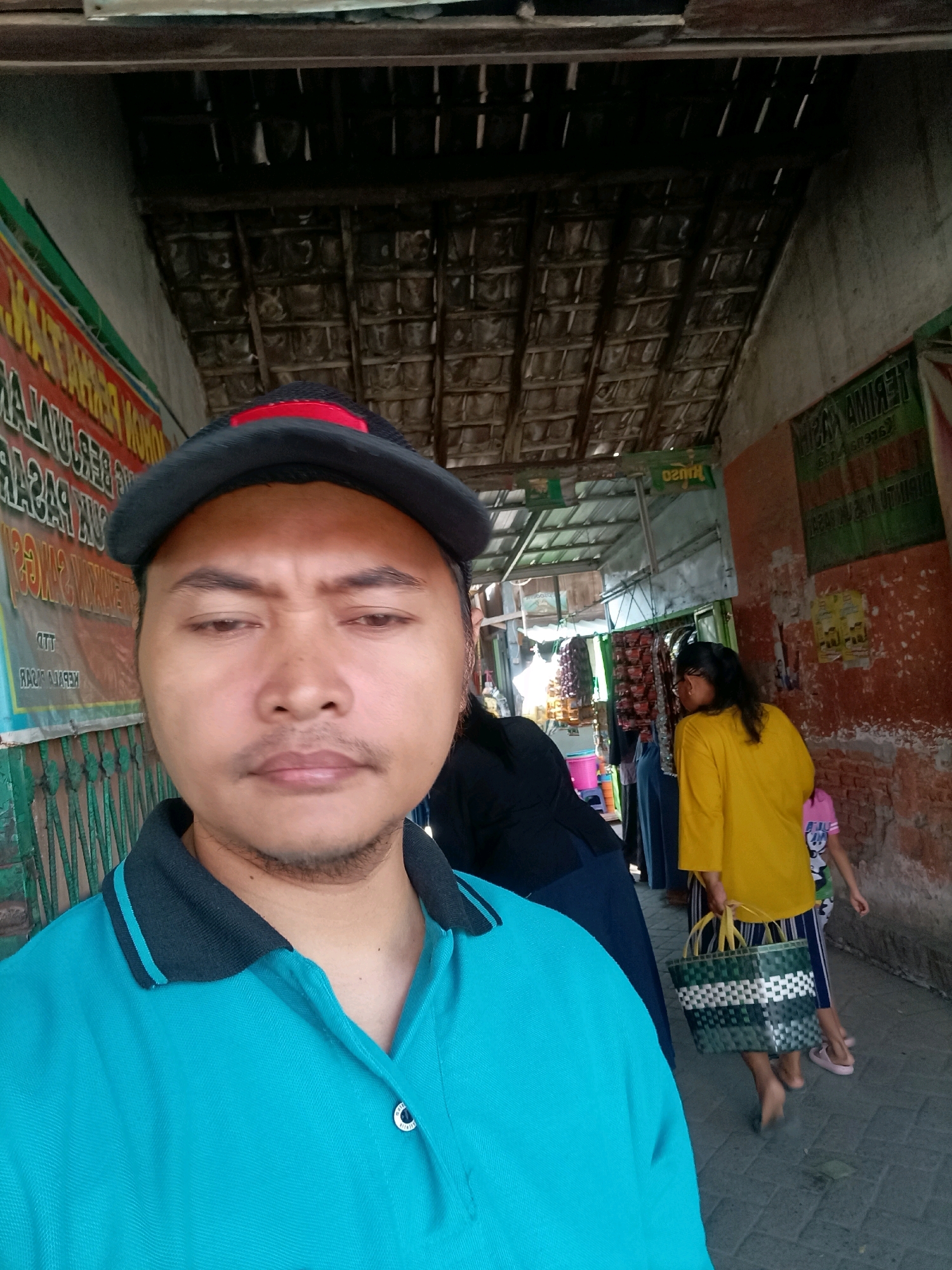 A man wearing a teal polo shirt and a dark baseball cap is taking a selfie in what appears to be a market alleyway in Badas, Indonesia. The roof above is constructed of thatched material supported by wooden beams. To the man's left, a poster with text is visible on a wall. In the background, other individuals are present. A woman in a yellow shirt is walking away, carrying a woven green and white bag. A young child in a pink shirt and blue pants is also visible behind the woman. The ground is paved with stone.