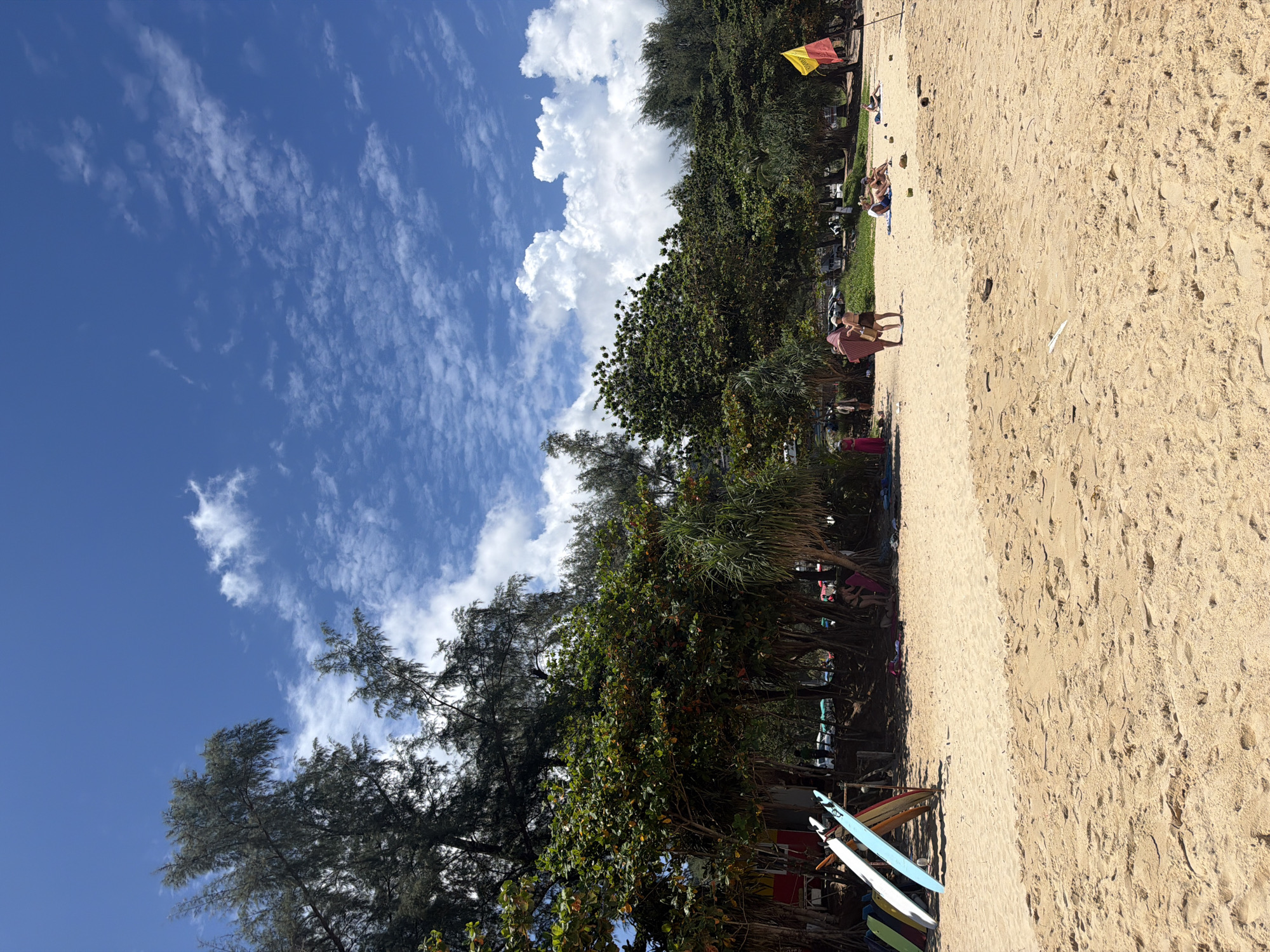 The image depicts a sunny day at a sandy beach, likely in Choeng Thale, Thailand, characterized by a clear blue sky with scattered white clouds. The foreground is dominated by a expanse of light brown sand, showing subtle undulations and footprints. To the left, several surfboards are propped upright, leaning against a wooden structure partially obscured by foliage.
In the midground, a line of lush green trees and palms forms a natural backdrop to the beach. Beneath the trees, a few individuals are visible. Two people, one in a bikini and the other in a light-colored outfit, are standing near the trees. Further to the right, several people are relaxing on the sand, some lying down on towels. A yellow and red flag is planted in the sand to the far right. The overall scene suggests a relaxed atmosphere typical of a tropical beach resort.