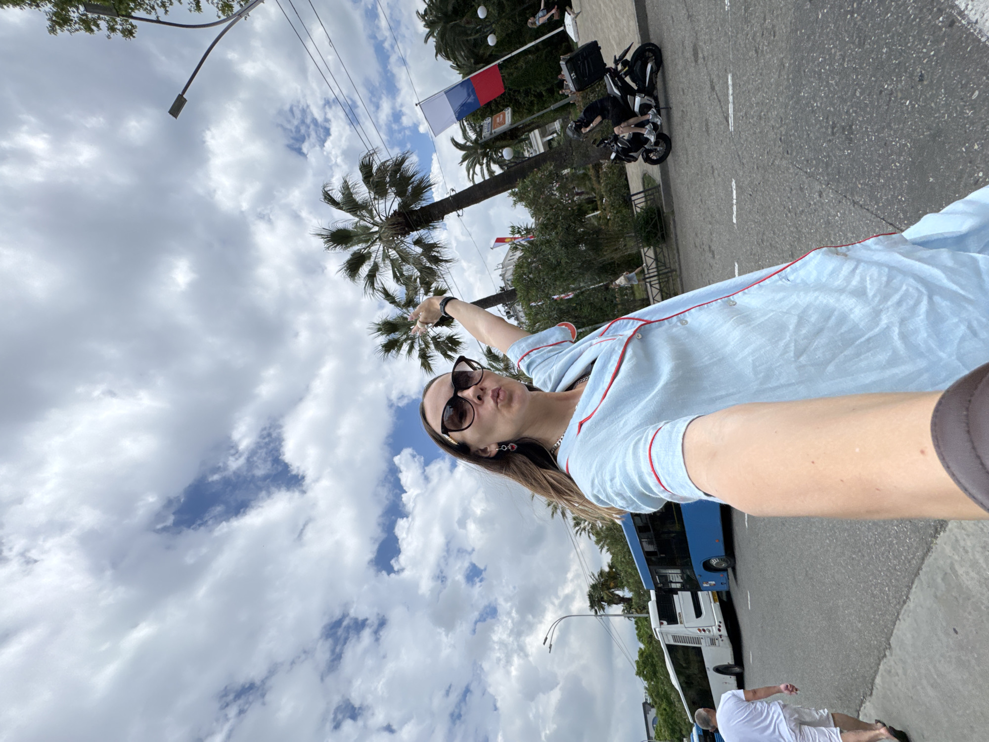 A woman with sunglasses and a light blue pajama set is posing for a selfie on a street in Sochi, Russia. She has her arm extended, holding the camera, and is pointing her other arm upwards. Behind her, palm trees and a cloudy sky are visible. Several buses are parked on the street, and a person on a motorcycle is also present. A Russian flag is displayed on a pole in the background. The woman is wearing heart-shaped earrings.