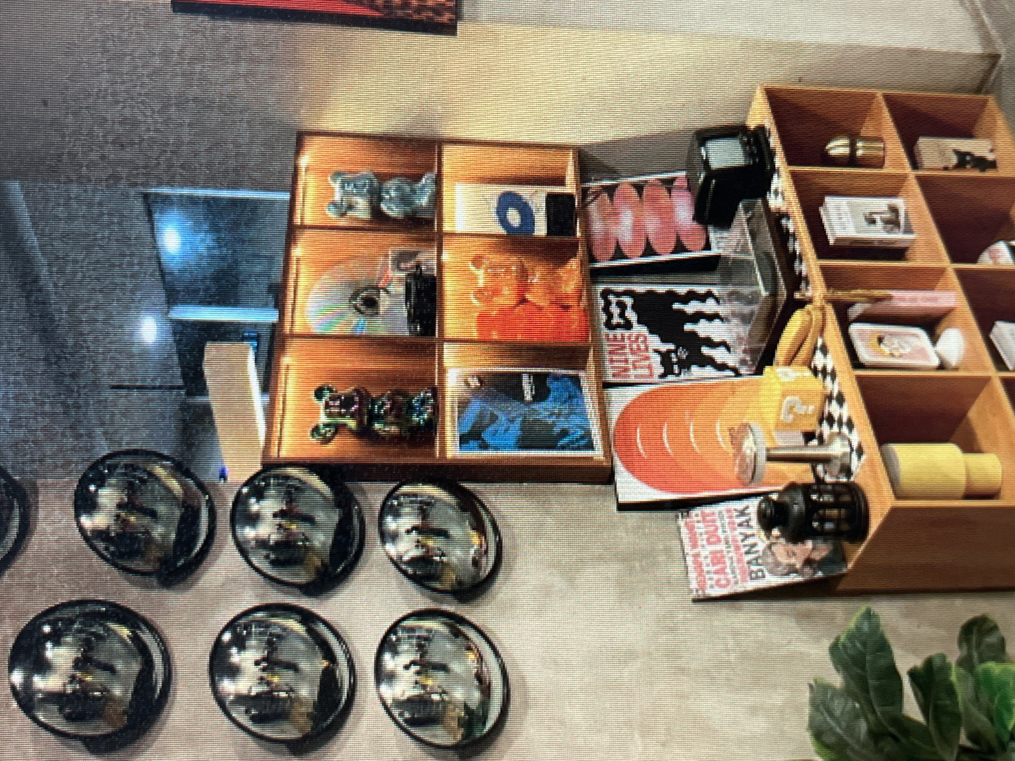 The image shows a section of a room, possibly a retail display or a home interior, featuring a wall adorned with reflective circular objects and a wooden shelf unit displaying various items. The reflective objects on the wall appear to be convex mirrors, each reflecting a dimly lit indoor scene with people. The wooden shelf unit, arranged in cubbies, holds decorative figures resembling stylized bears, what looks like a vinyl record player, a small vintage-style television, books, and framed artwork or posters. The overall aesthetic suggests a modern, curated space with an emphasis on collectibles and artistic display. The background wall has a textured, concrete-like appearance. The location of Sejahtera, Indonesia is not directly discernible from the visual content of the image itself.