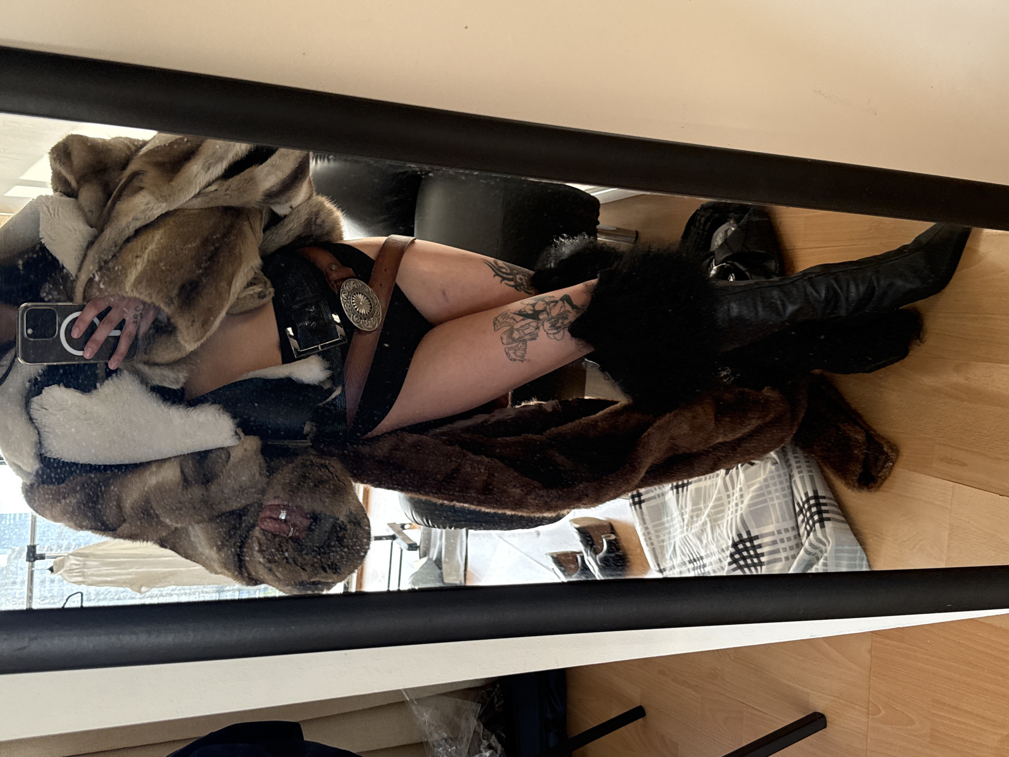 A person is captured in a mirror selfie, seen from approximately the waist down, though the image is initially oriented sideways. The individual is wearing a voluminous, multi-toned fur coat, featuring shades of brown, white, and darker patches. Beneath the coat, a black garment, possibly underwear or a bodysuit, is visible. A wide brown leather belt with a large, ornate circular silver buckle (concho style) cinches the waist.
The person's legs are bare and display multiple black tattoos, including what appears to be a floral or intricate design on the thigh. Black, furry leg coverings or boots extend down into black leather boots. An iPhone with a clear case, showcasing a prominent circular MagSafe magnet, is held in the person's left hand, with a ring visible on one finger.
The reflected background reveals a light wooden floor, a dark piece of furniture (possibly a sofa or chair), and a grey, white, and black plaid blanket. Through a distant window, an urban cityscape with buildings is visible, suggesting an elevated view in an urban environment like London, United Kingdom. The overall scene indicates an indoor setting during daylight hours, given the visible light from outside. No specific text is legible in the image.